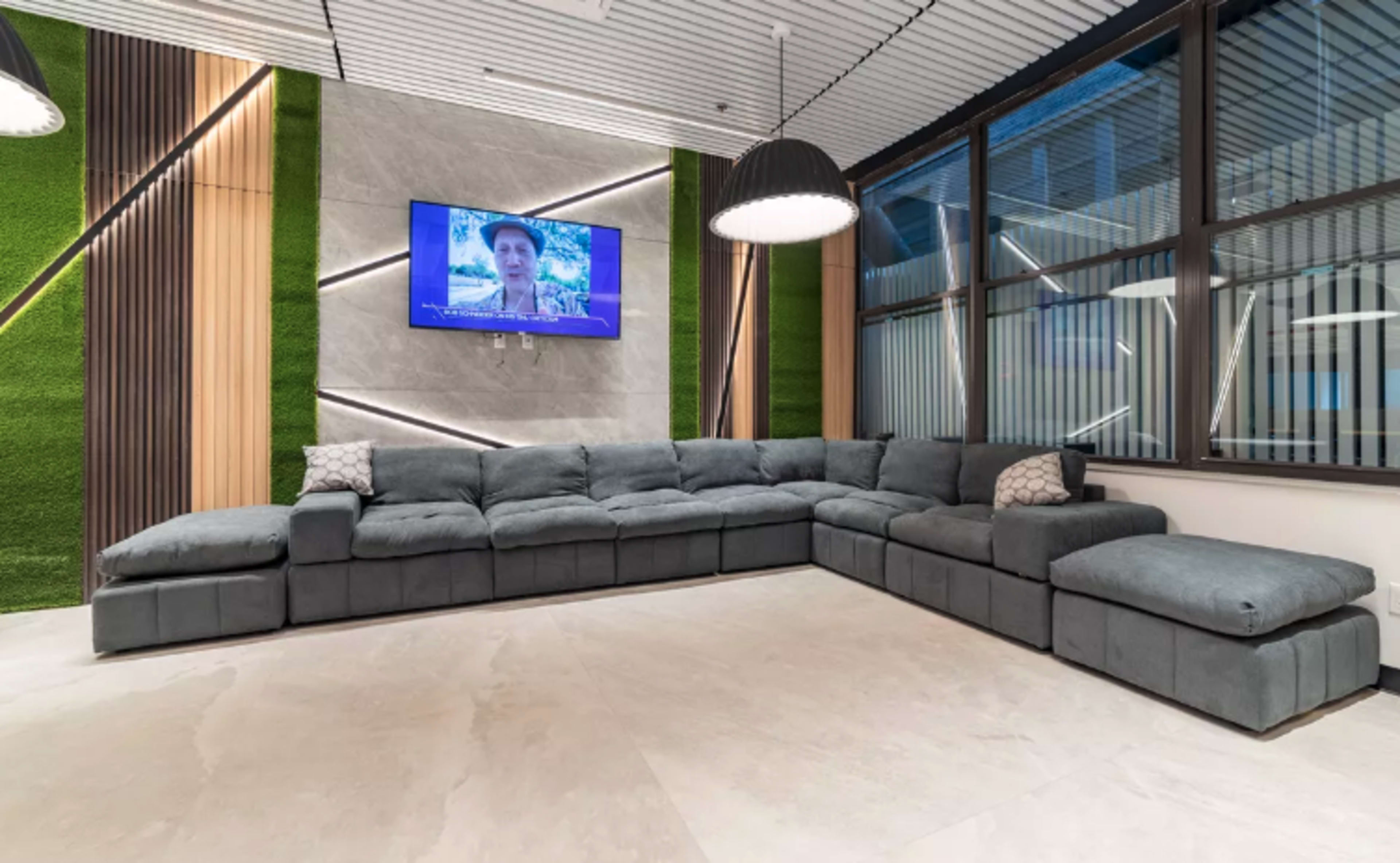 A modern, L-shaped sectional sofa arranged in a bright room with large windows and a wall-mounted television.