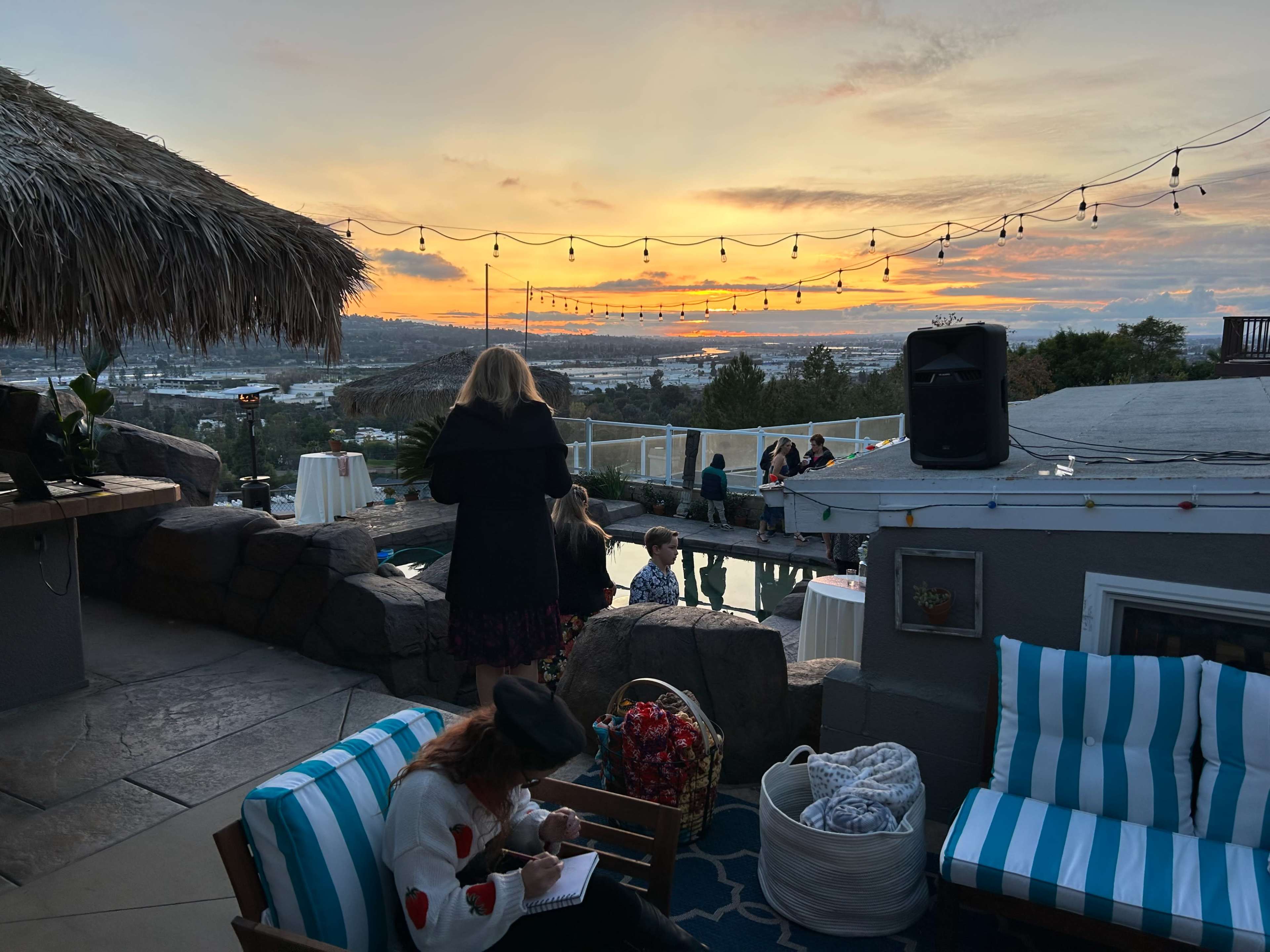 A group of people gathers on a patio overlooking a sunset, with string lights illuminated above and a pool in the foreground.