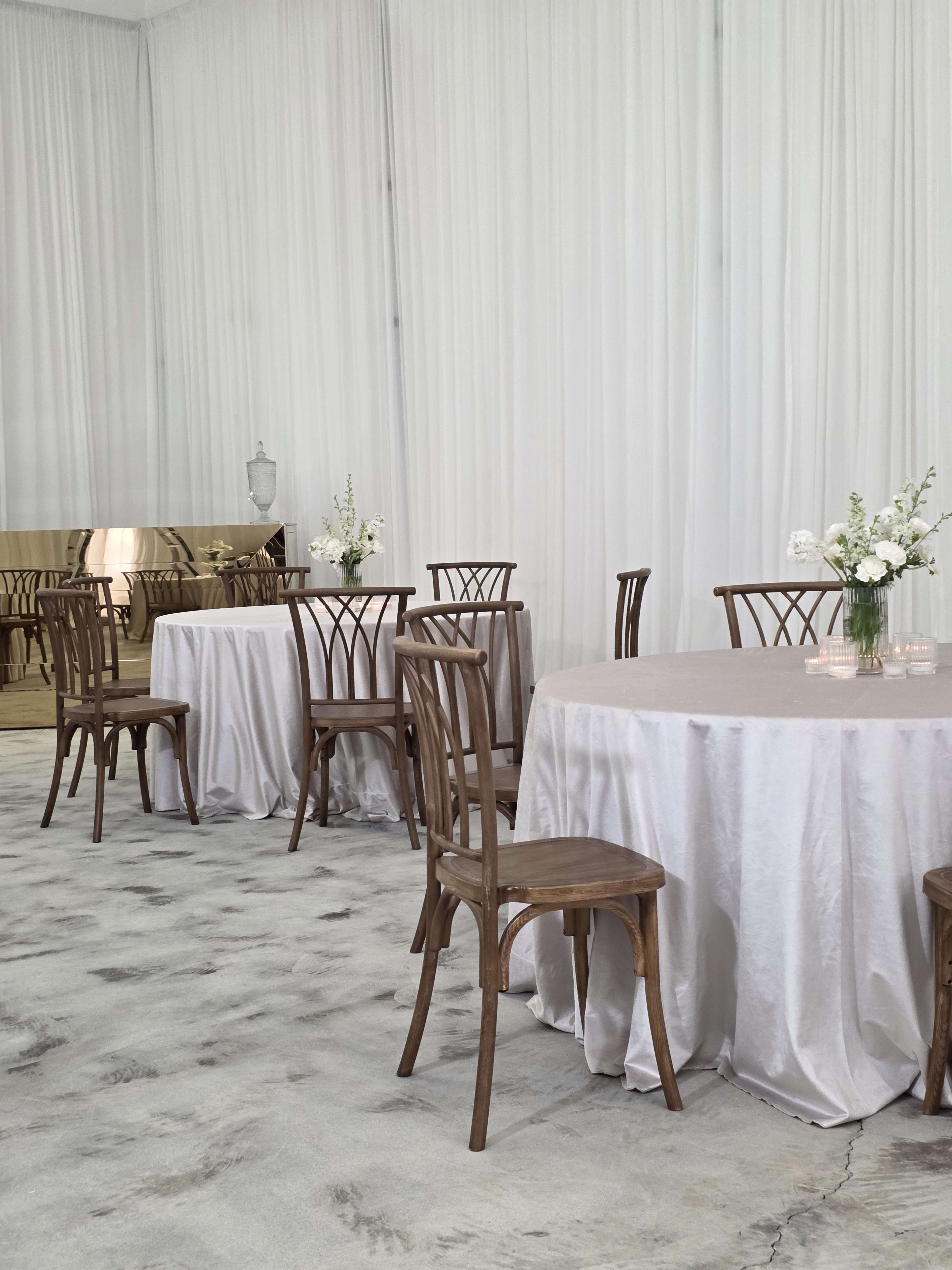 The image shows a minimalist indoor event space featuring several wooden chairs and tables covered with light-colored tablecloths, accompanied by floral centerpieces.