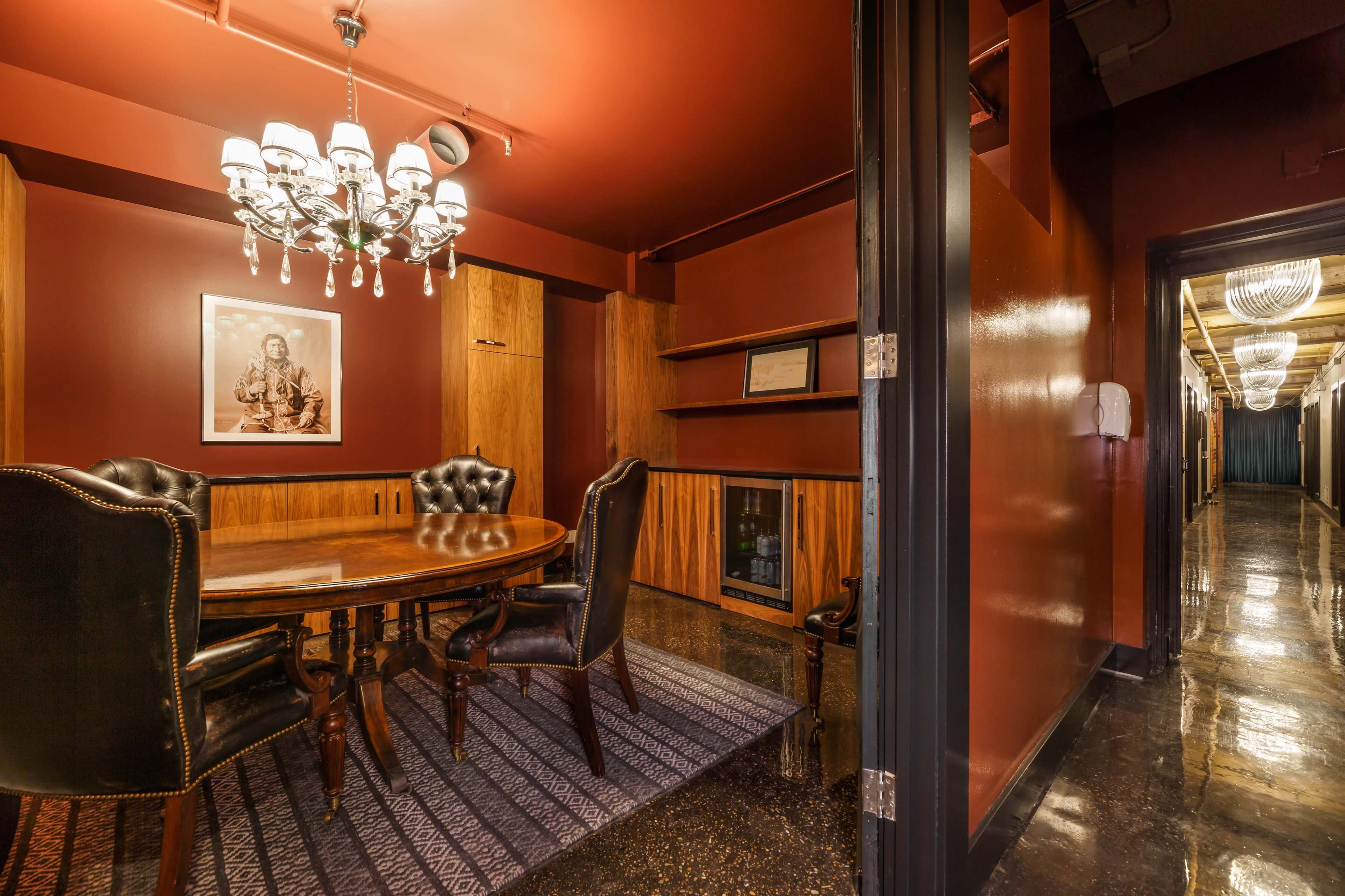 A conference room features a round wooden table surrounded by high-backed chairs, with a chandelier overhead and dark red walls.