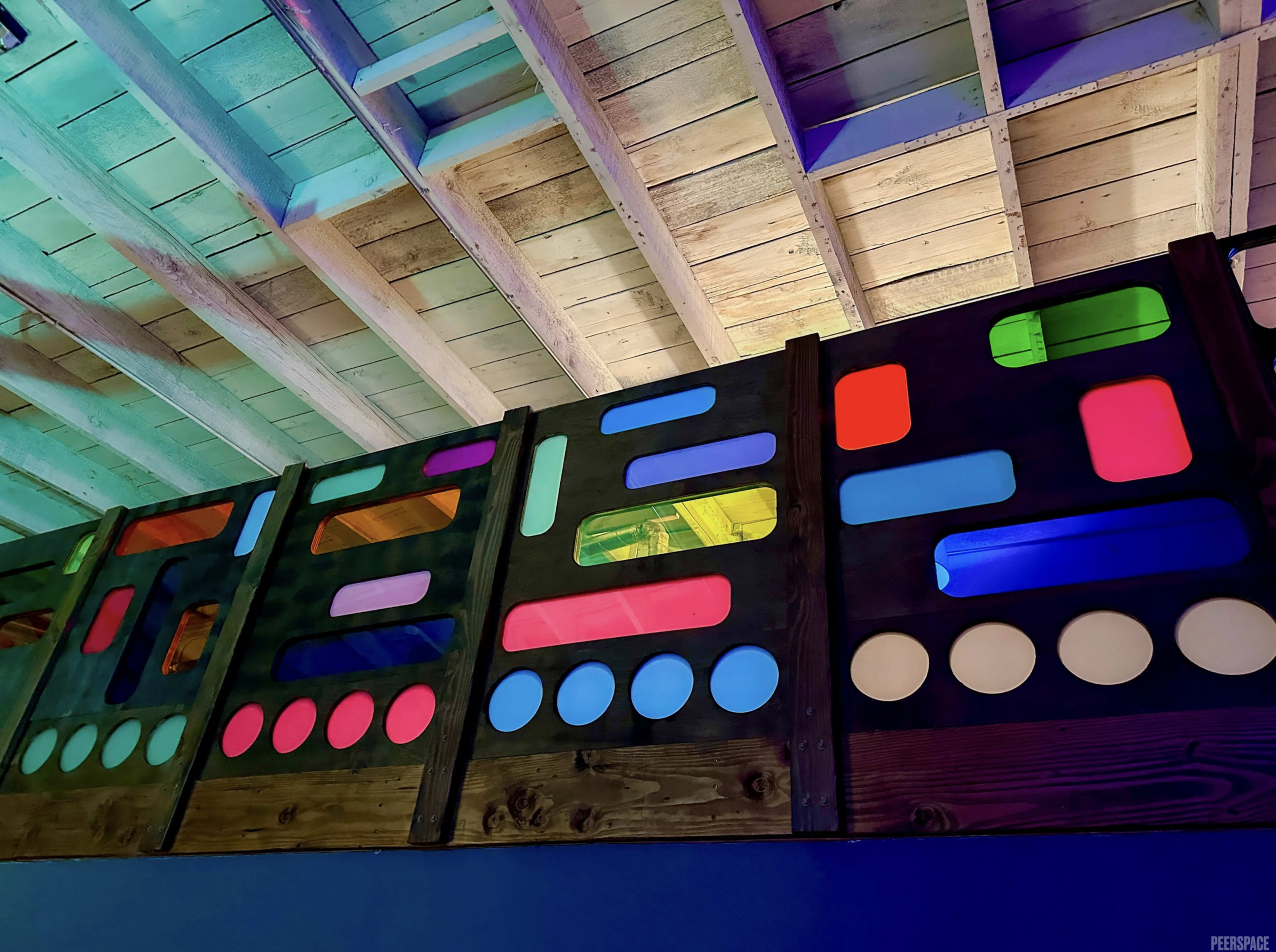 The image features a wooden structure with a brightly lit, colorful design consisting of various geometric shapes and patterns against a ceiling of exposed beams.