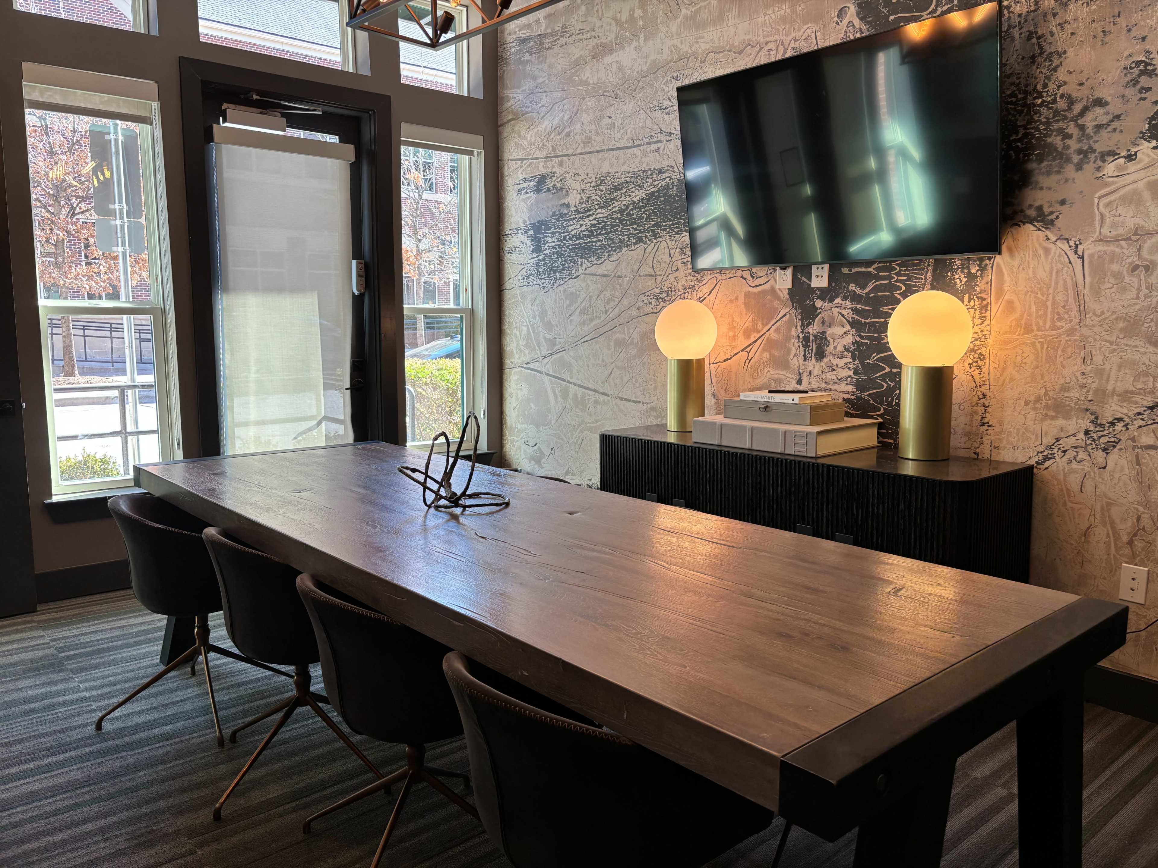 A large wooden conference table is set in a meeting room with a television mounted on the wall and two round lamps on a side console.