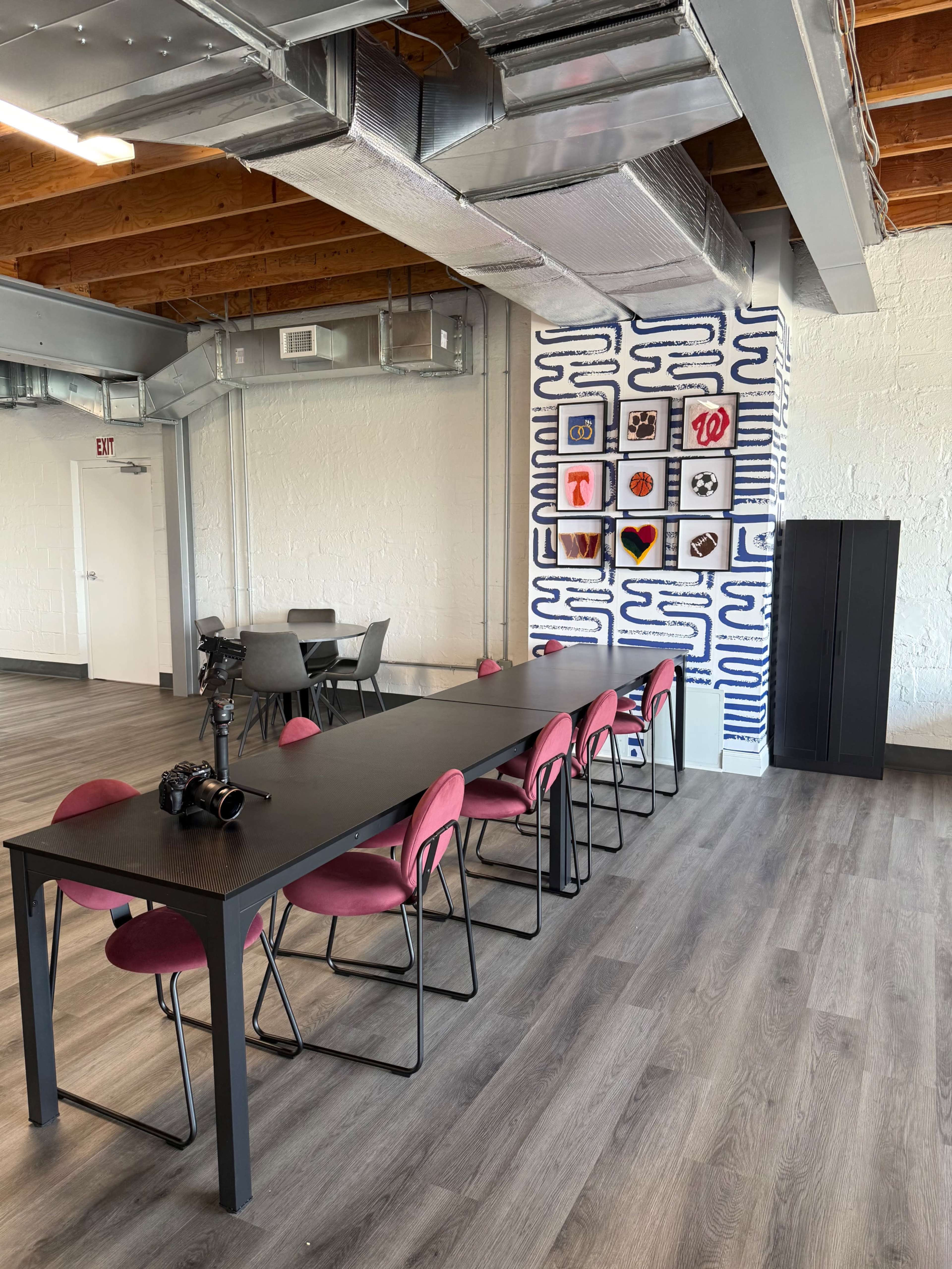A spacious, modern workspace features a long black table surrounded by pink chairs, with a wall displaying framed artwork and a camera on the table.