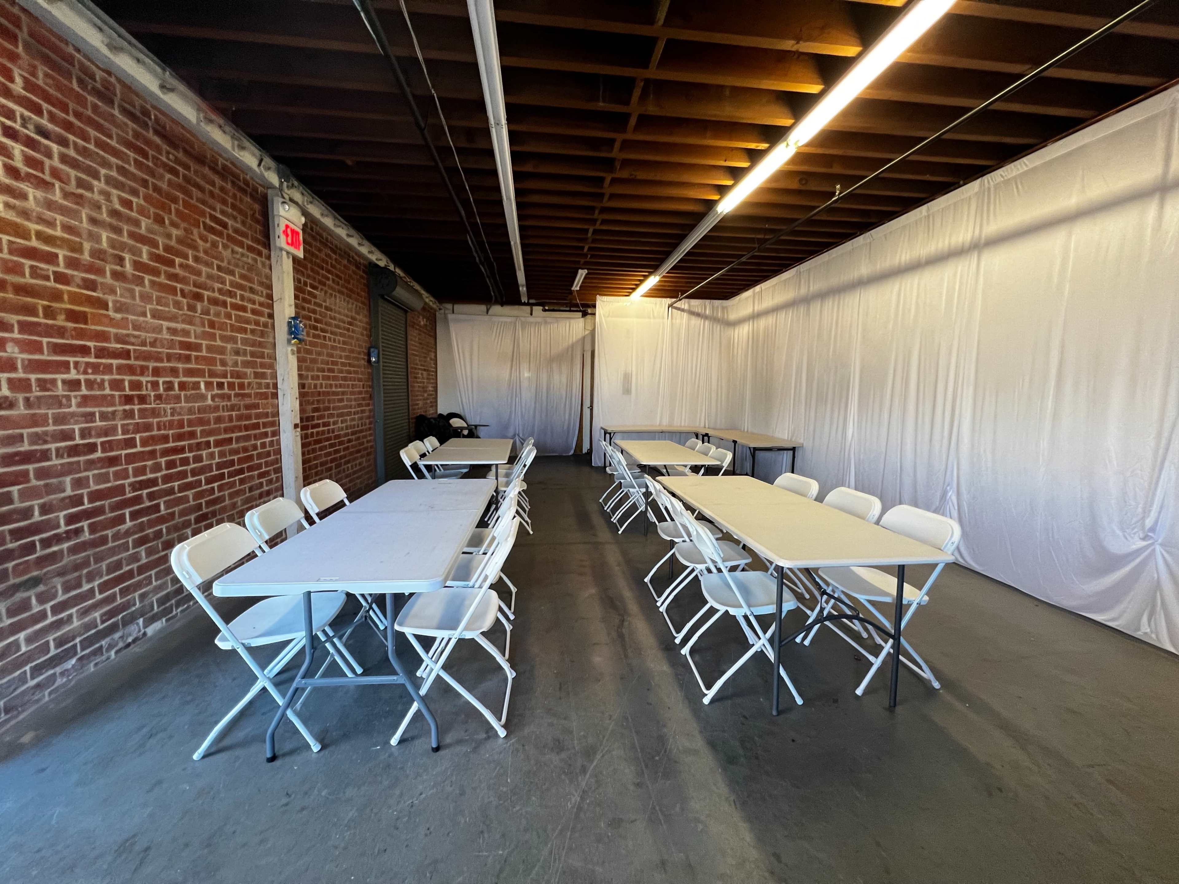 A spacious room with white tablecloths covering several folding tables arranged in two parallel rows, against a backdrop of exposed brick walls.