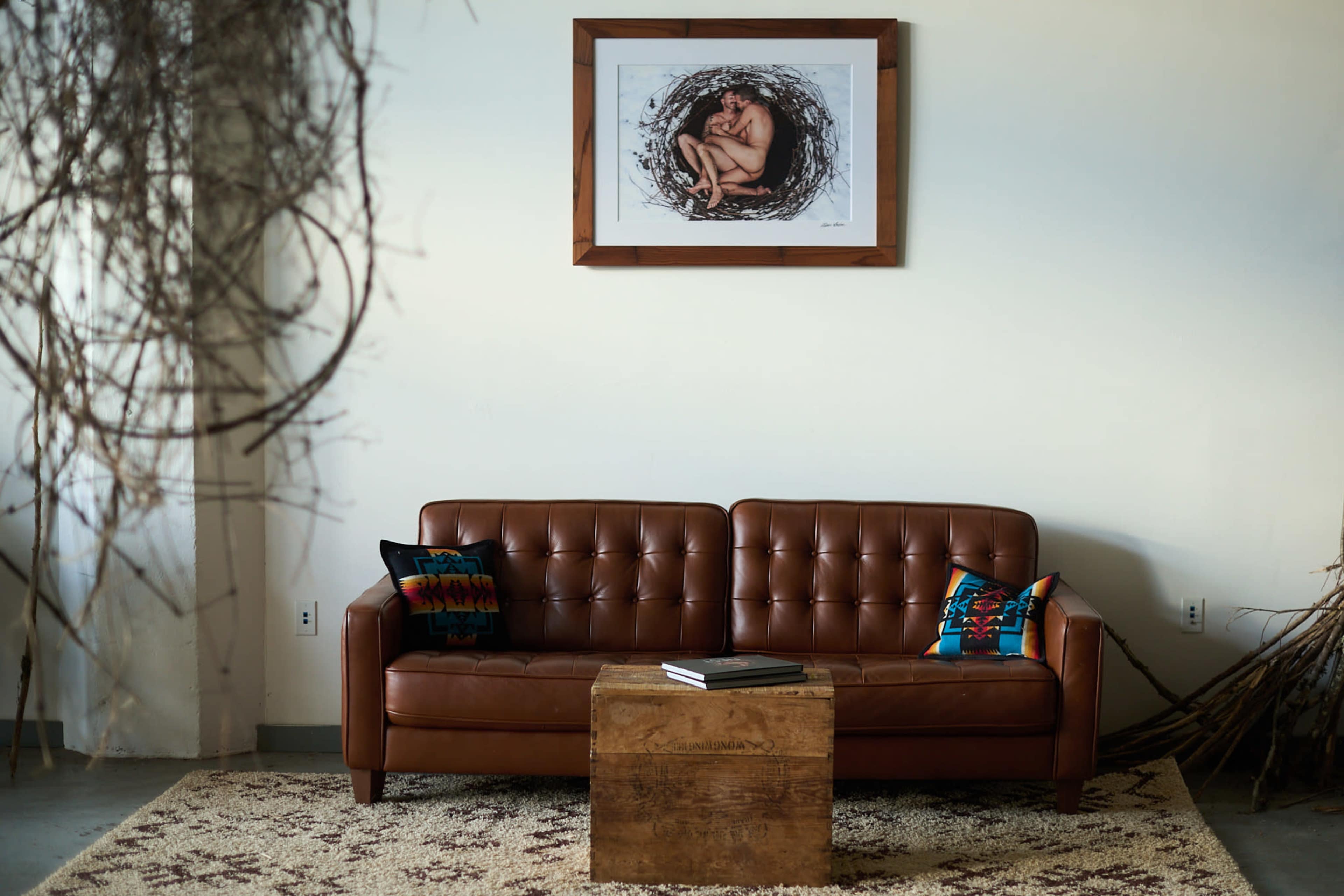 The image features a brown leather sofa with colorful pillows, a wooden coffee table, and an art piece hanging above against a plain wall.