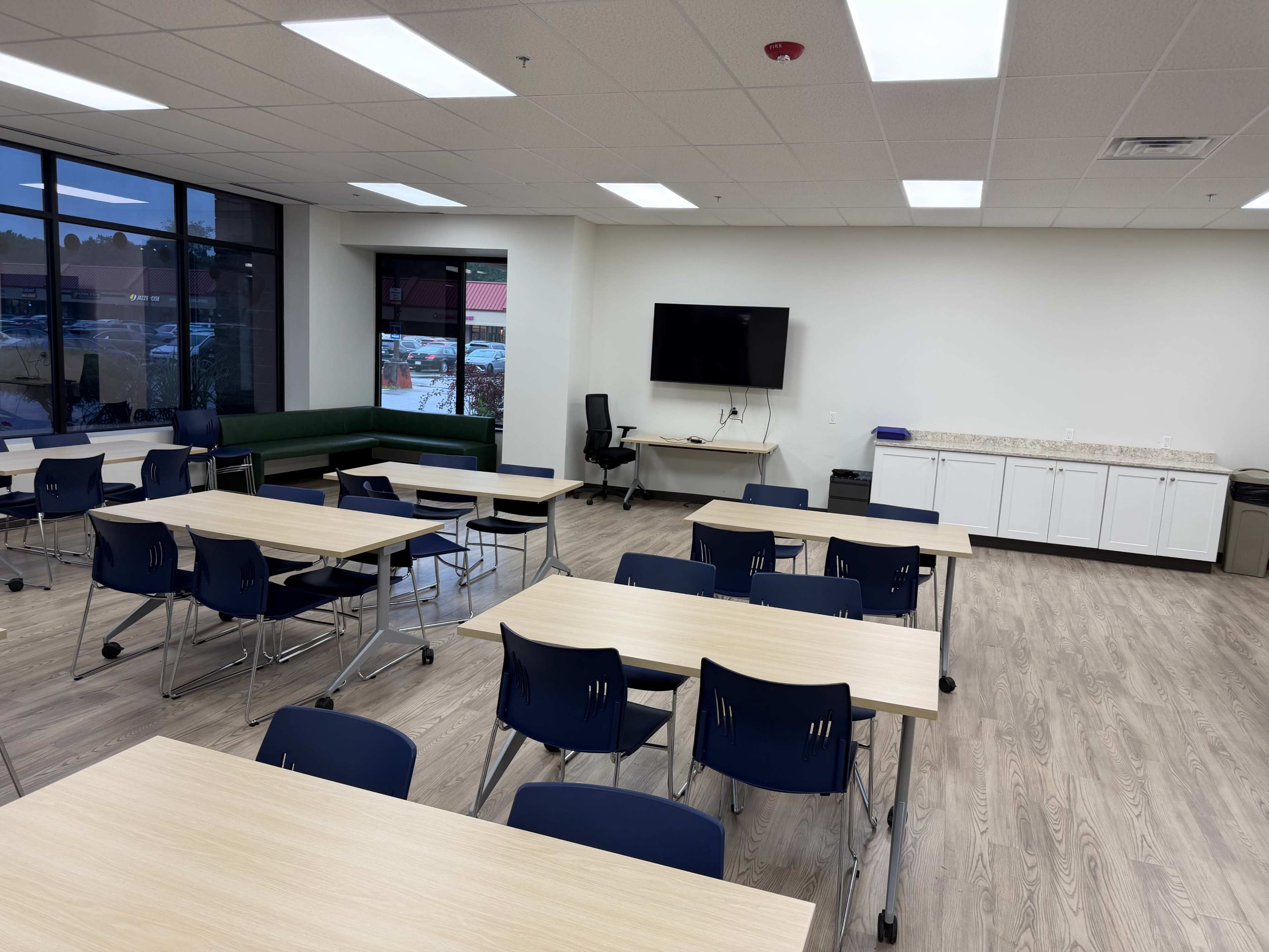 The image shows a spacious classroom or meeting area with multiple tables and chairs arranged for use, along with a television mounted on the wall and a counter with storage cabinets.