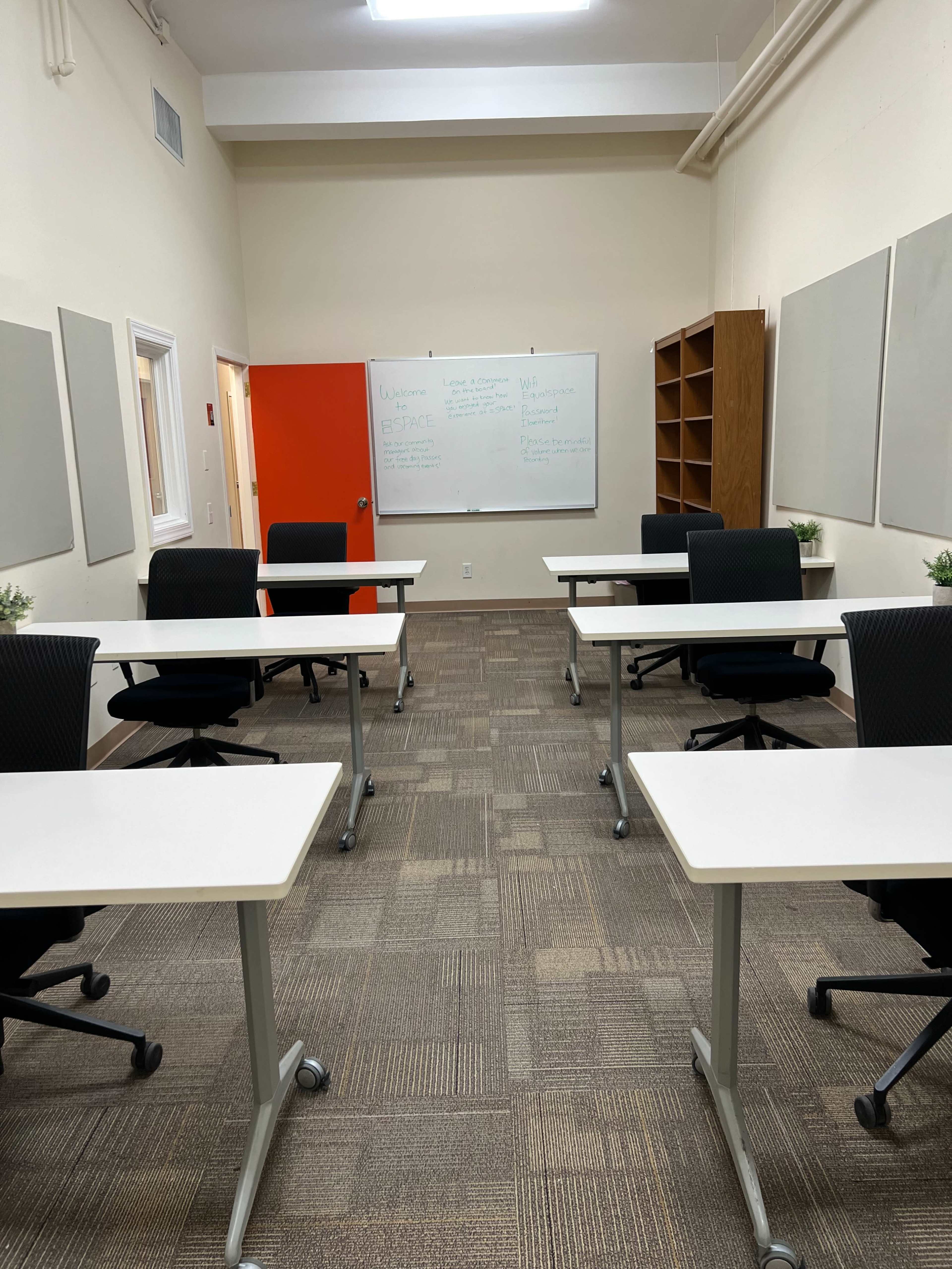 A classroom with several white tables and black chairs arranged around the room, featuring a whiteboard and a red door.