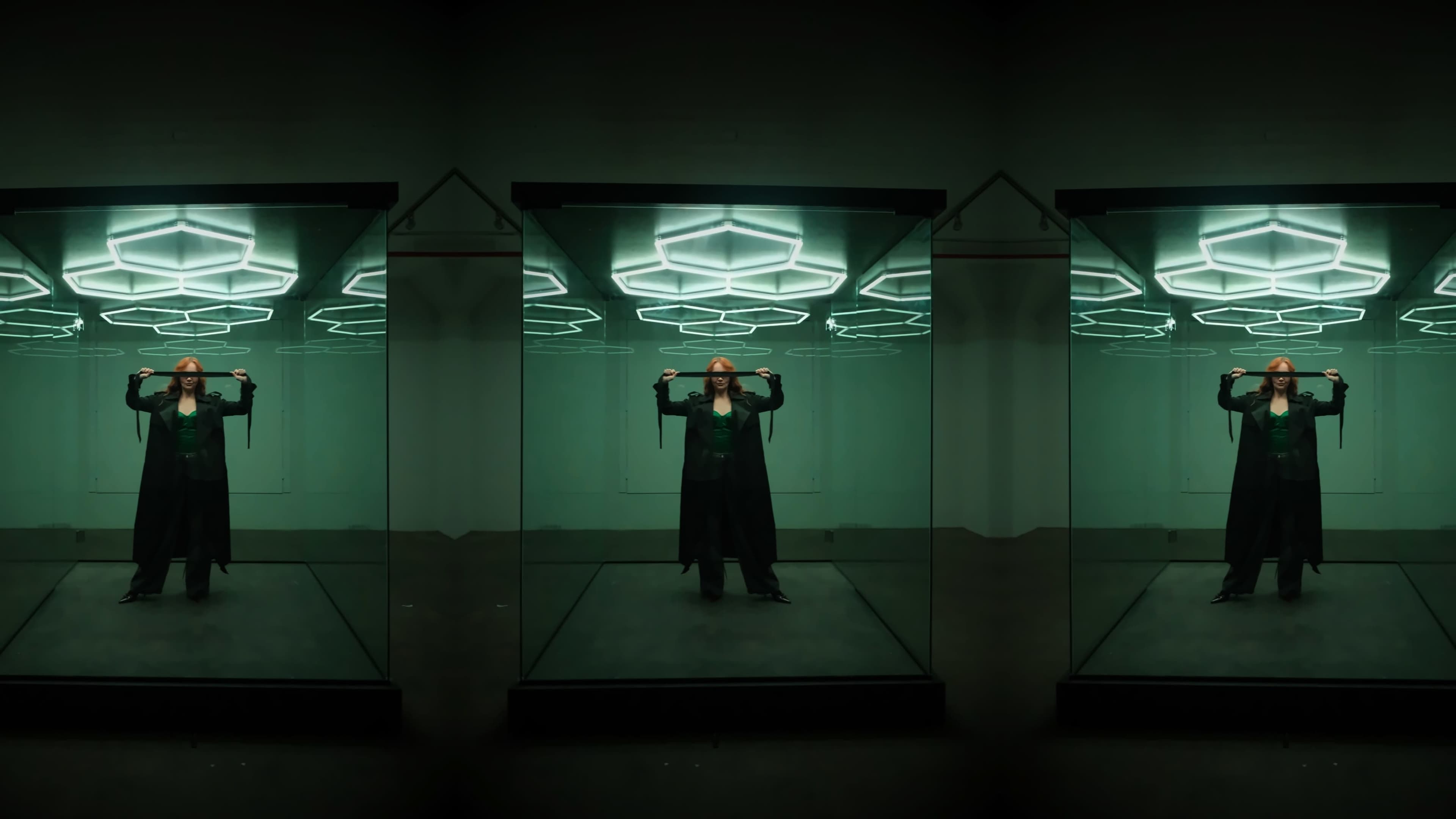 A person in a black cape stands confidently in a mirrored room with hexagonal lights overhead.