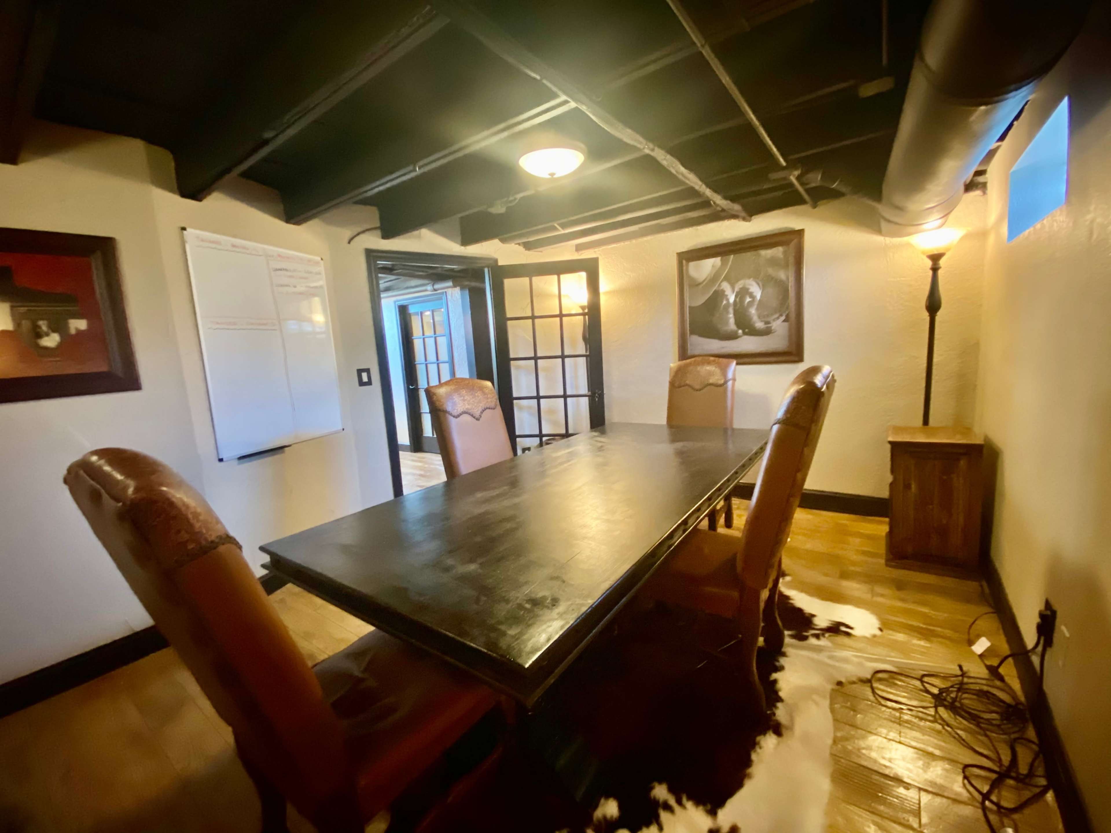 A conference room features a dark wooden table surrounded by six chairs, with a wall-mounted whiteboard and a lamp in the corner.
