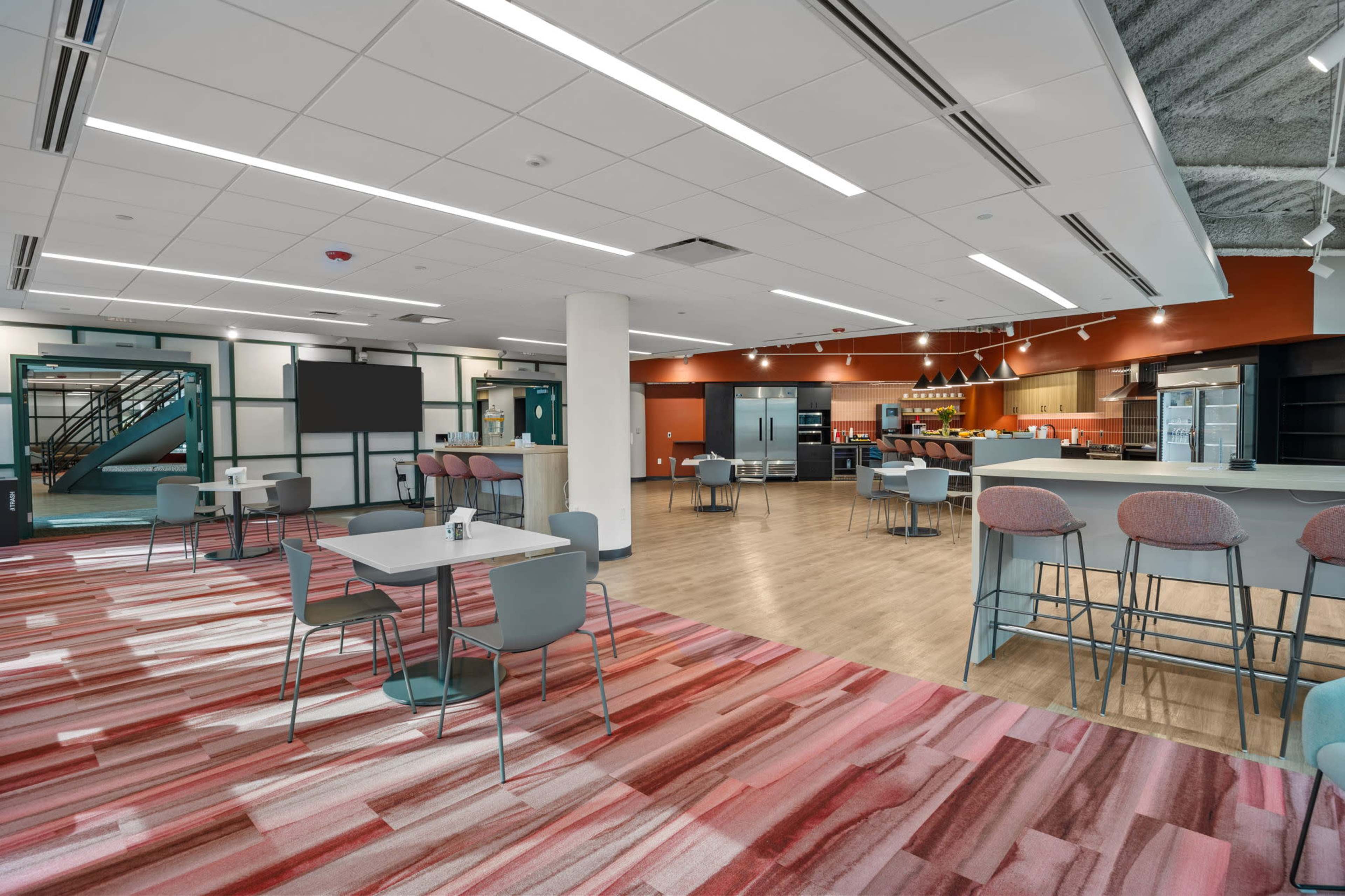 A modern, open-plan common area with tables, chairs, and a kitchenette in the background.