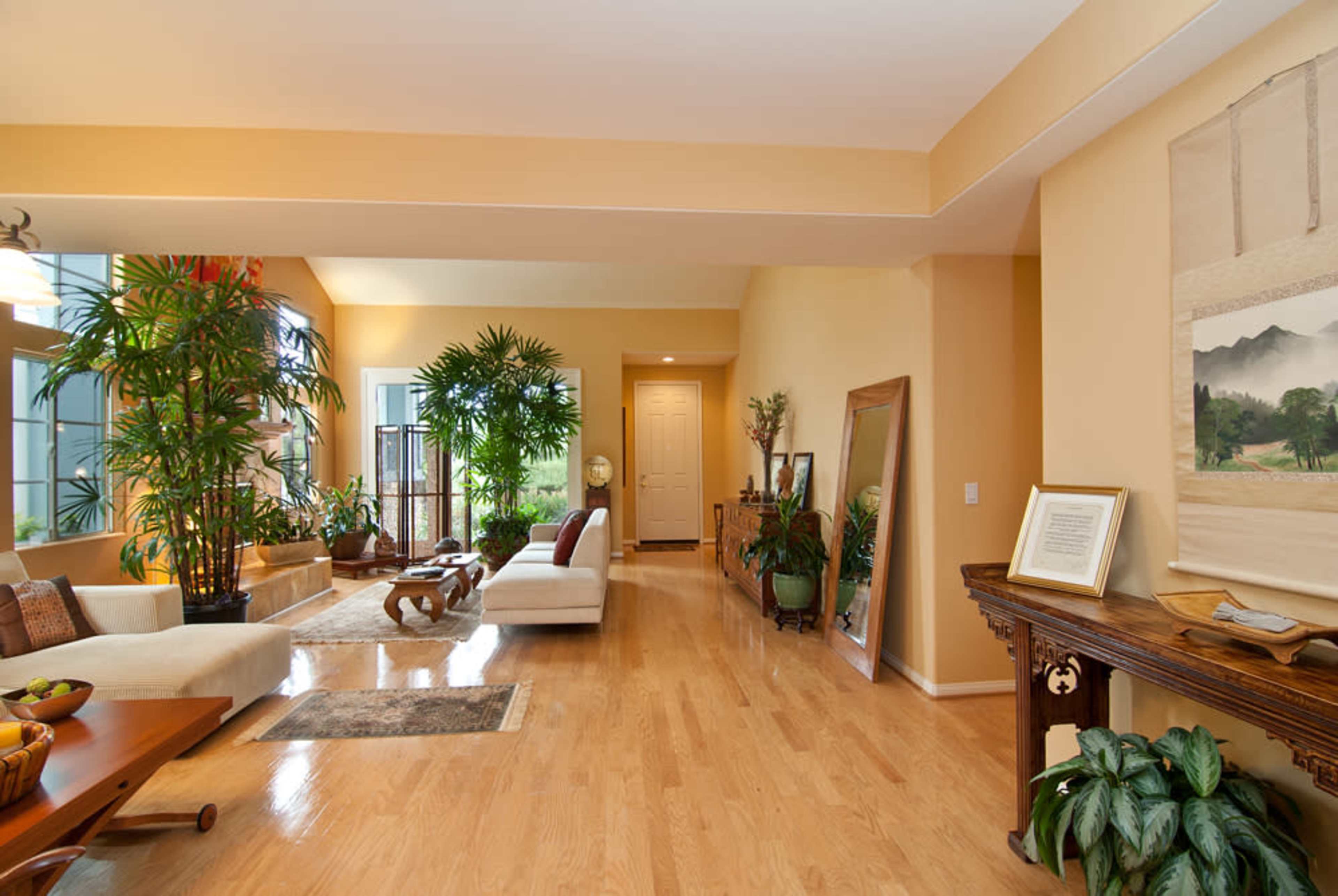 A spacious living room filled with plants, featuring a light-colored sofa, wooden furniture, and large windows that allow natural light to enter.
