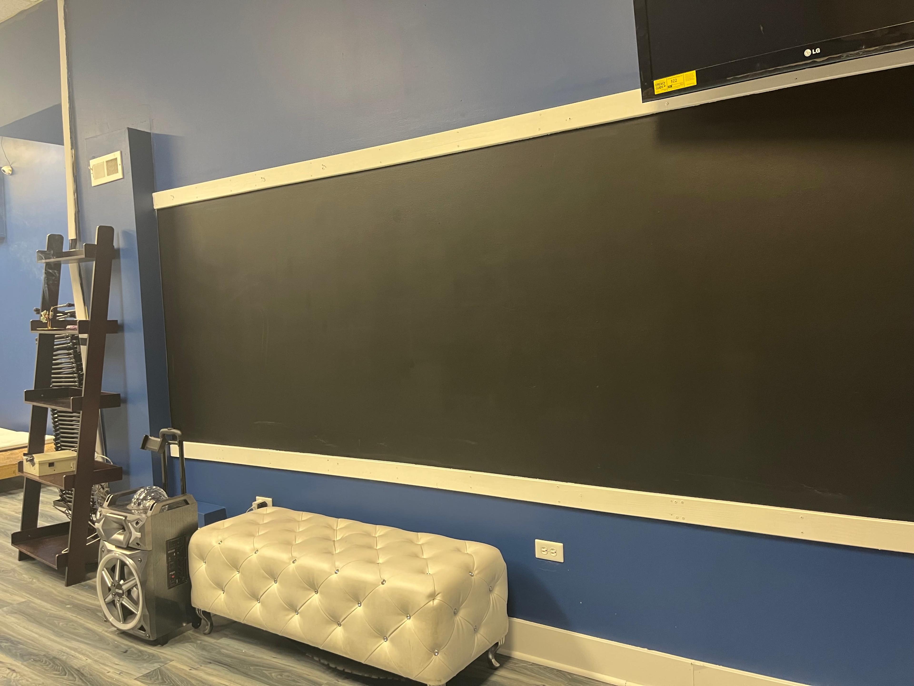 A wall-mounted black chalkboard beside a tufted bench and a television mounted above it in a blue room.
