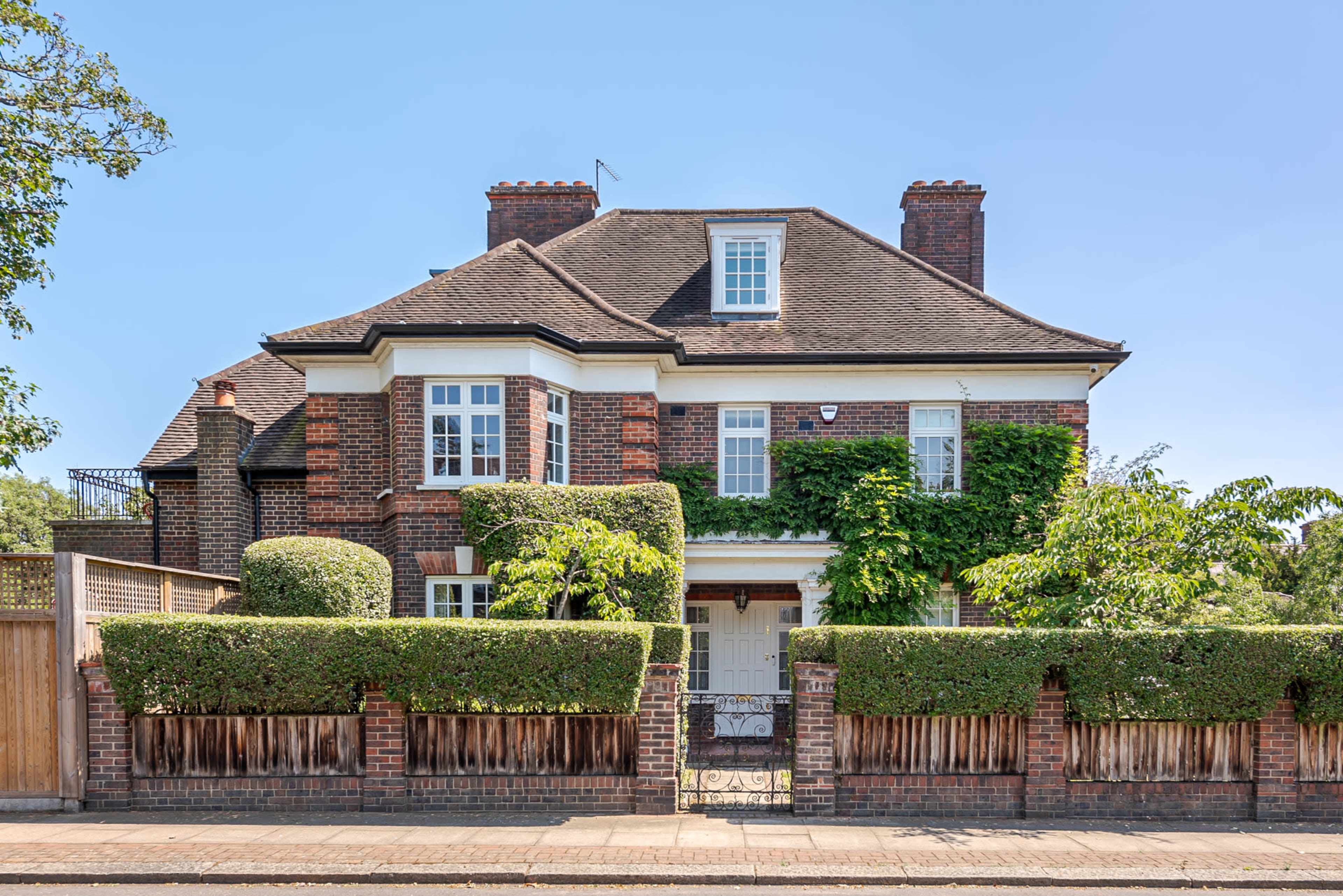 A large brick house with a steep roof and neatly trimmed hedges is set behind a wooden fence on a quiet street.