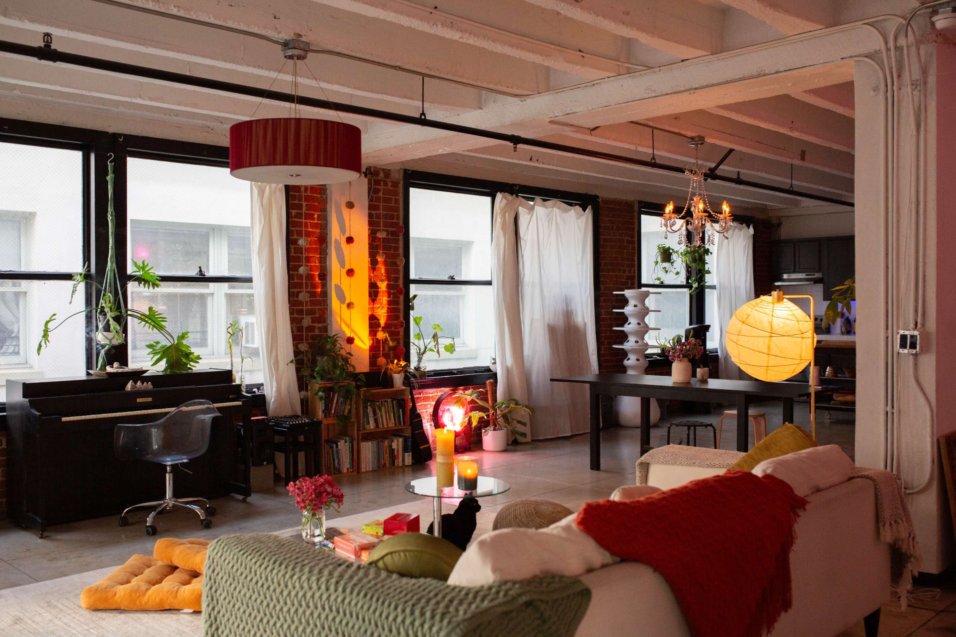 The image shows a modern living space with large windows, a piano, a dining table, and various plants, illuminated by warm lighting.