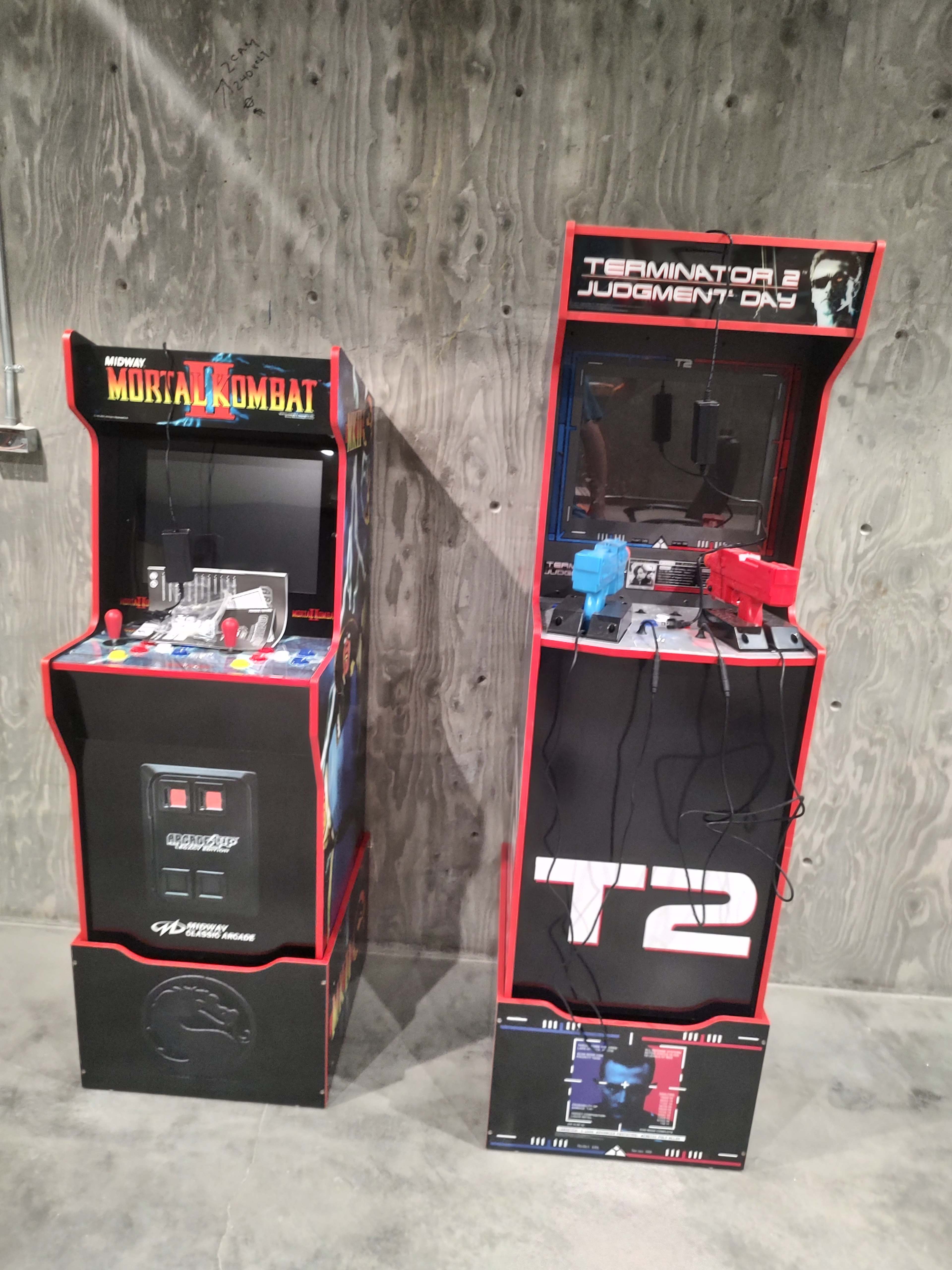 Two arcade game machines: one for "Mortal Kombat" on the left and another for "Terminator 2: Judgment Day" on the right, both set against a concrete wall.