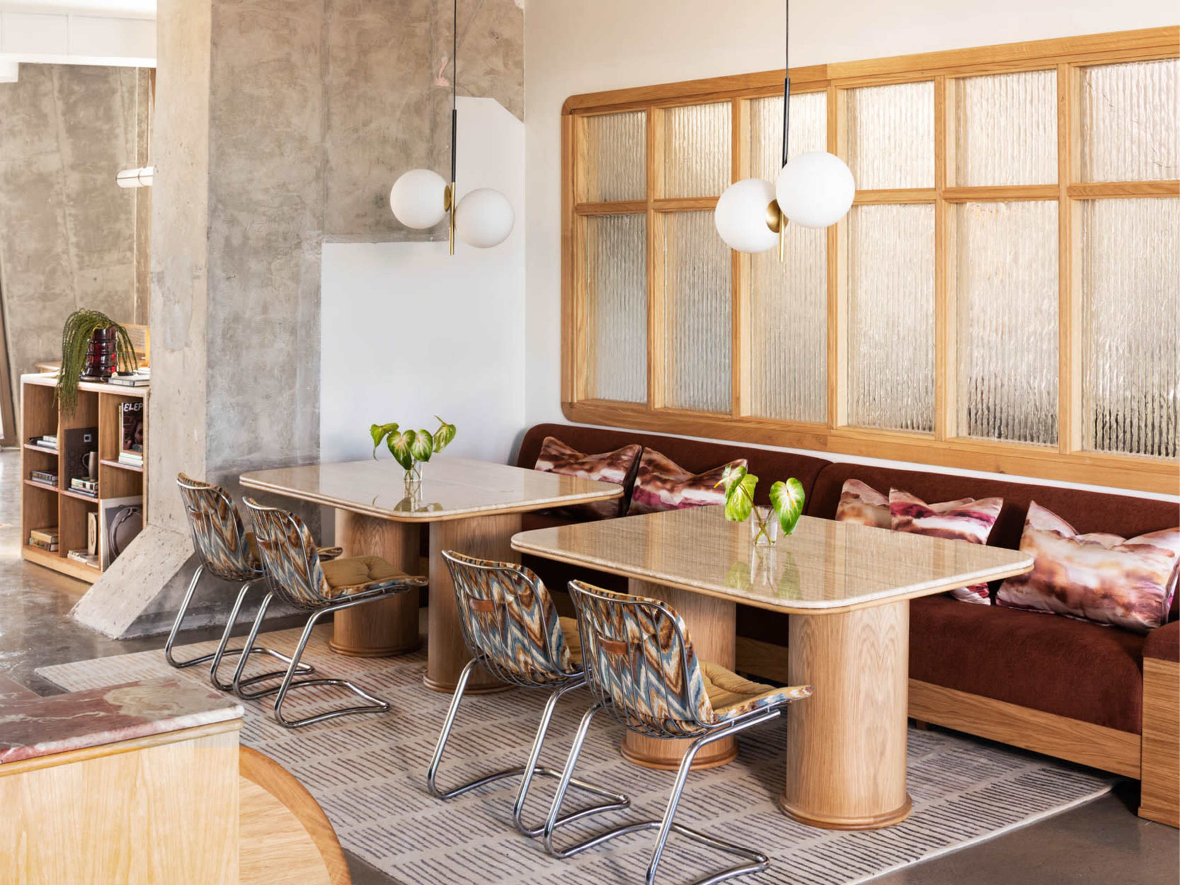 The image shows a modern interior space featuring two wooden tables surrounded by chairs, a textured wall, and a cozy seating area with cushions.