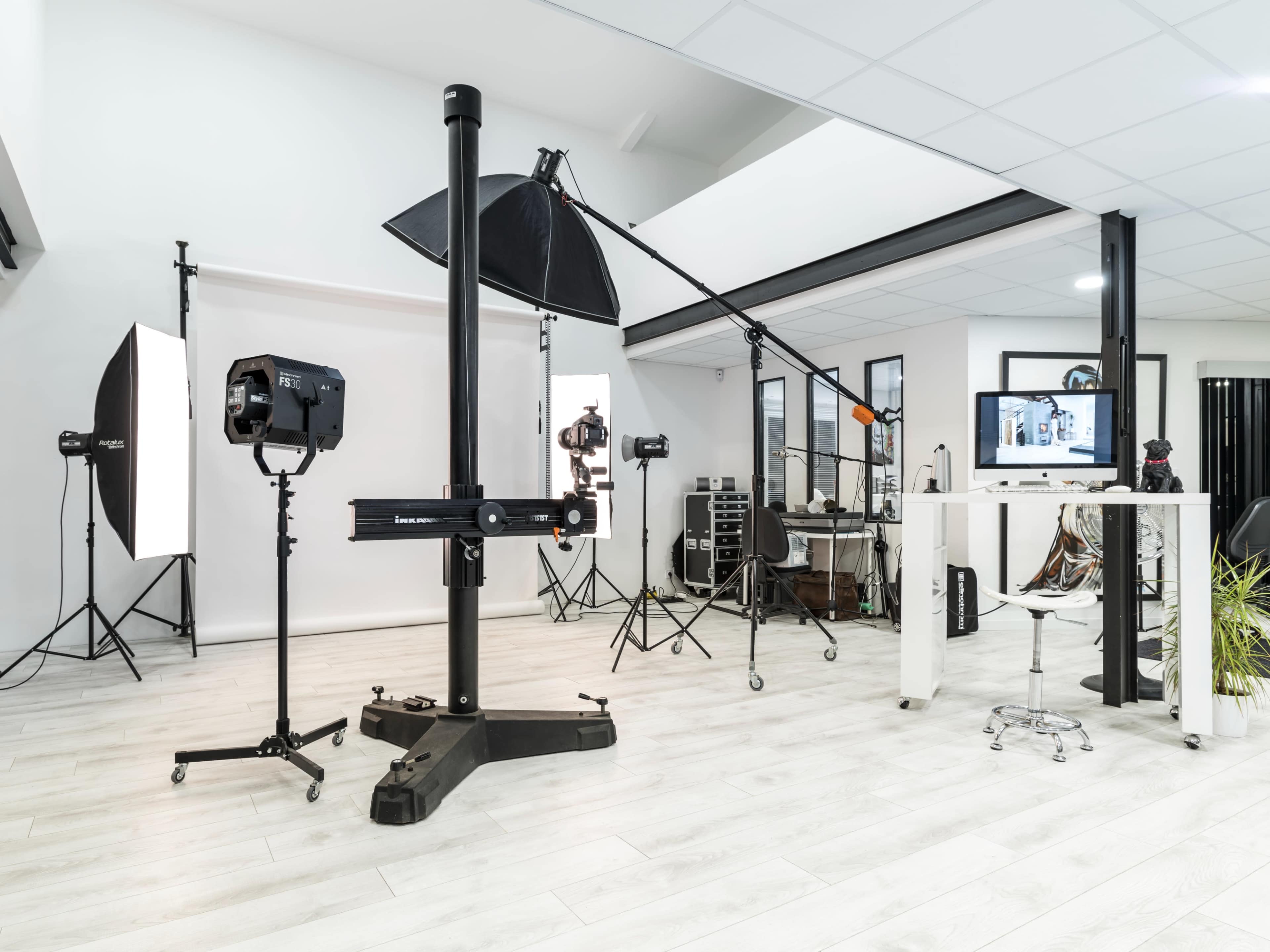 The image depicts a well-equipped photography studio featuring various lighting equipment, a backdrop stand, and a workspace with computer and seating.