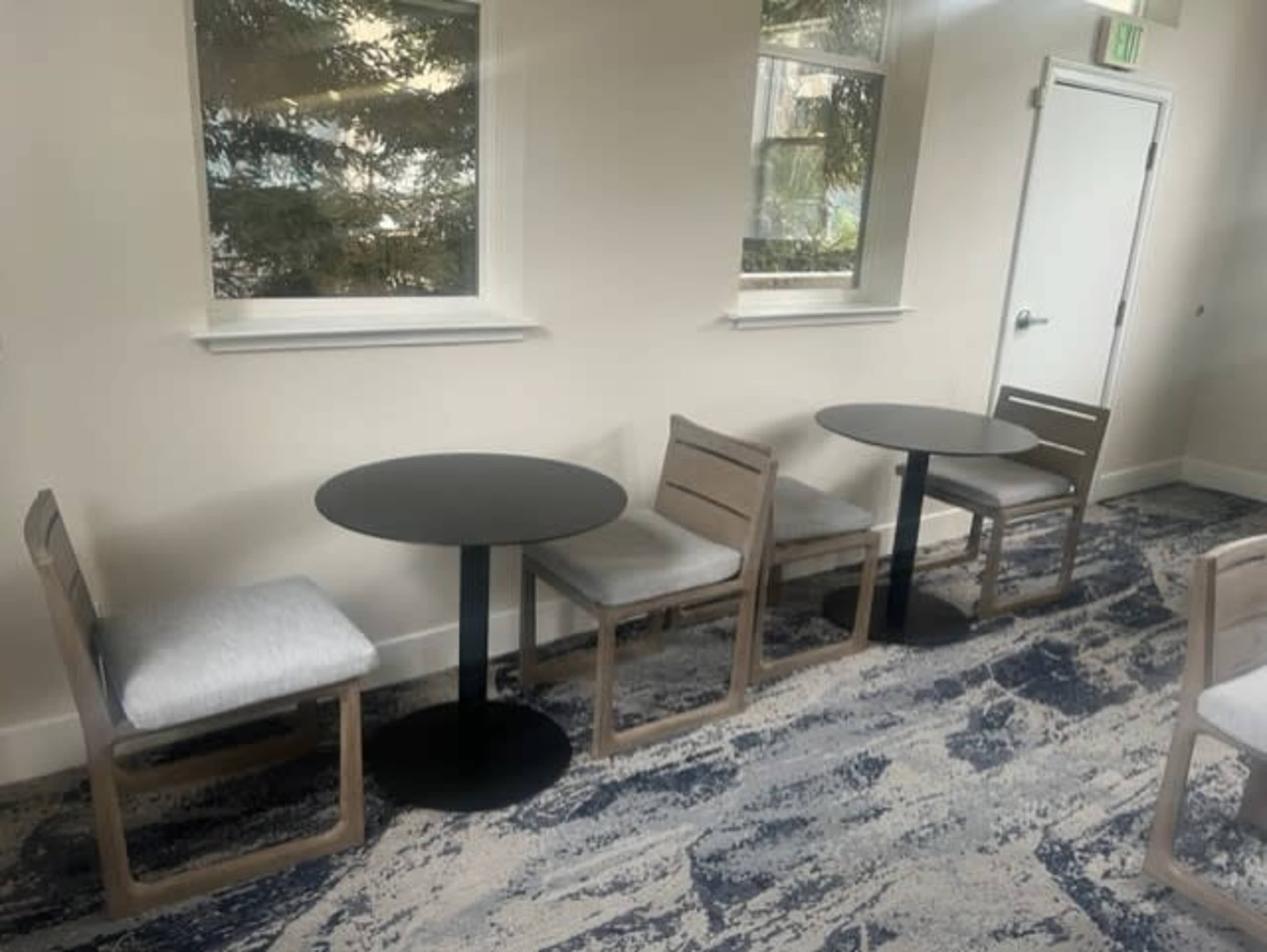 A small area with two round tables and four chairs, situated on a patterned carpet beneath large windows.