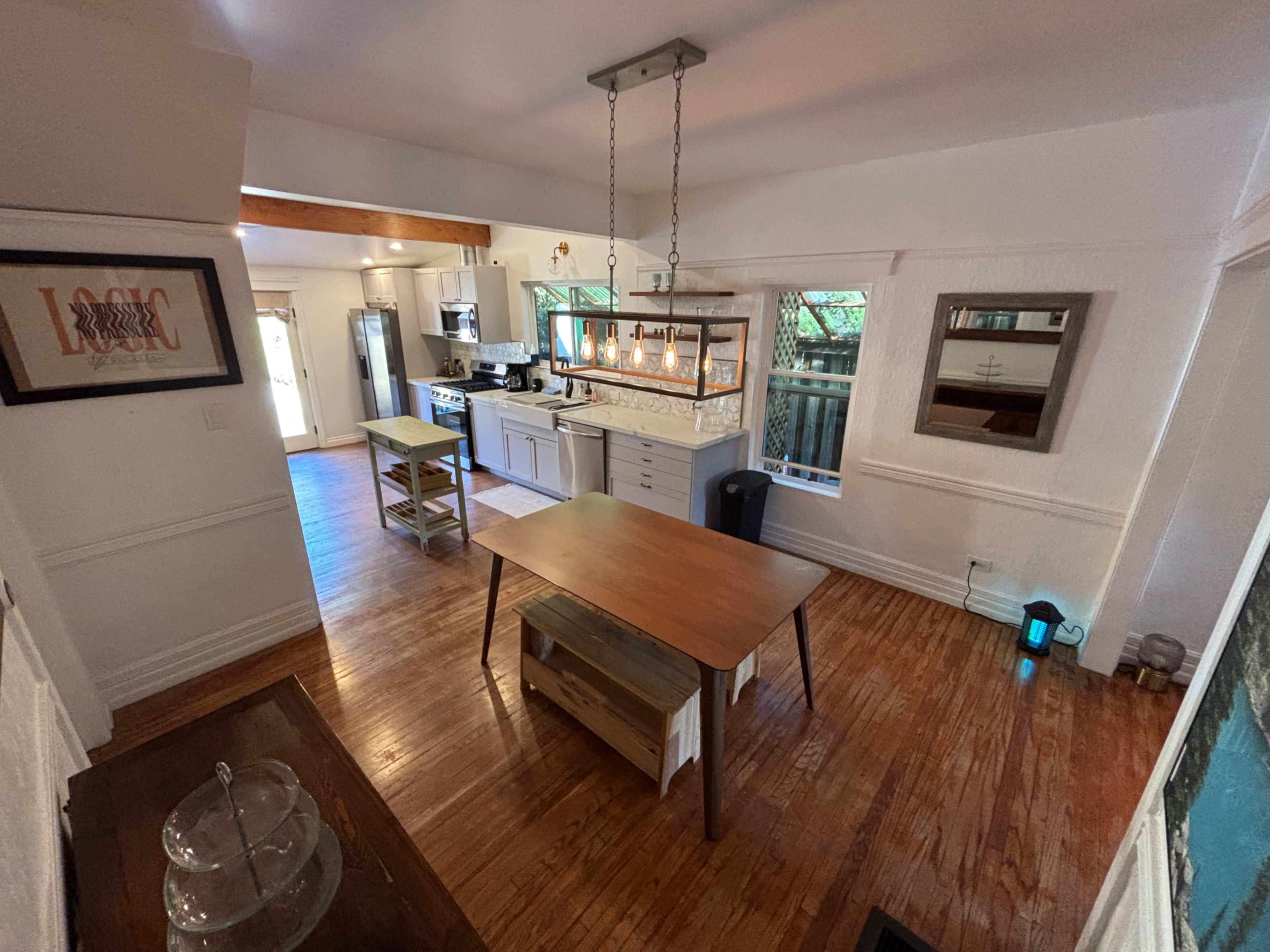The image shows a bright, modern kitchen and dining area with hardwood floors, a wooden table in the center, and a chandelier above it.