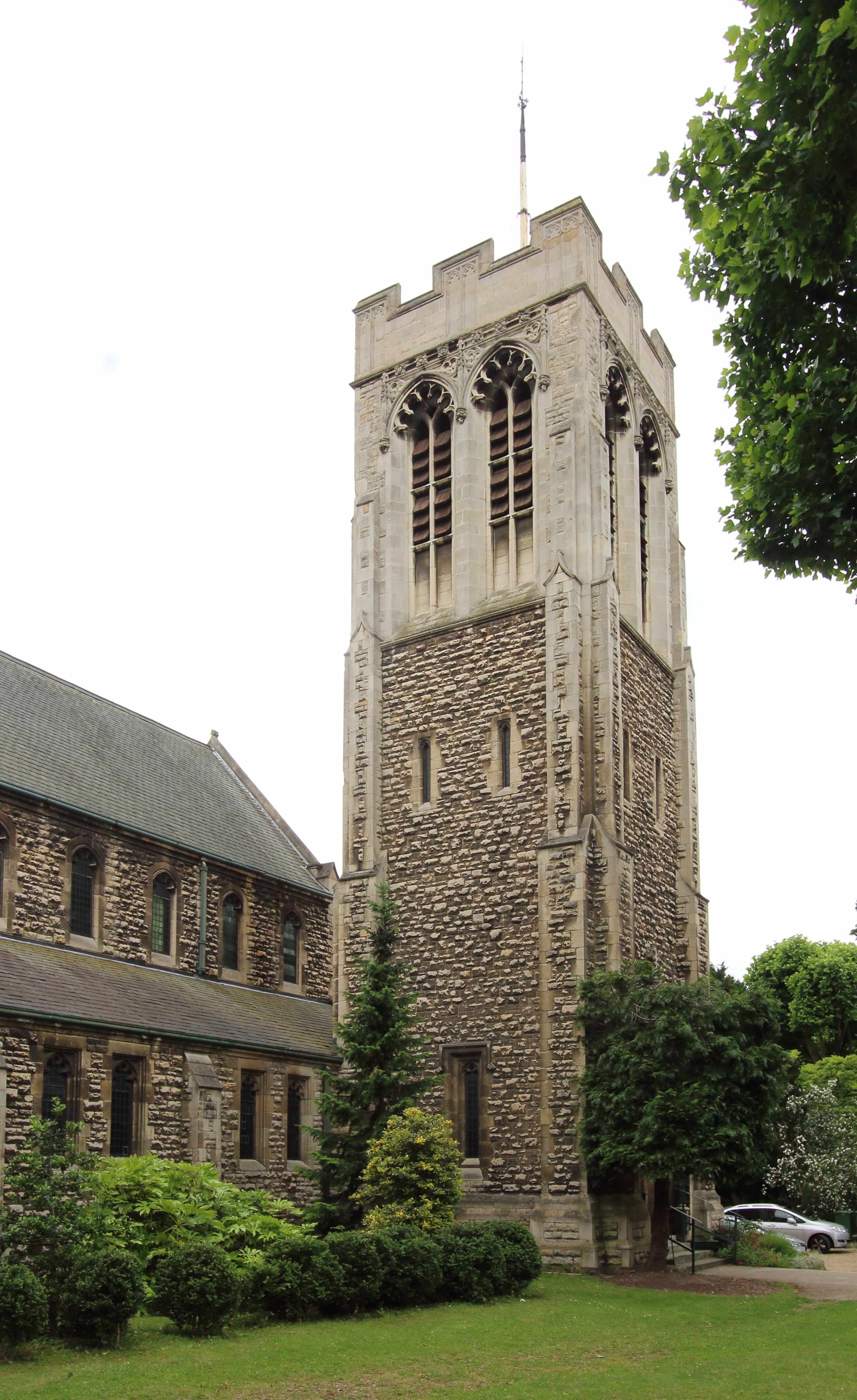 A stone church tower with large windows next to a building, surrounded by greenery.