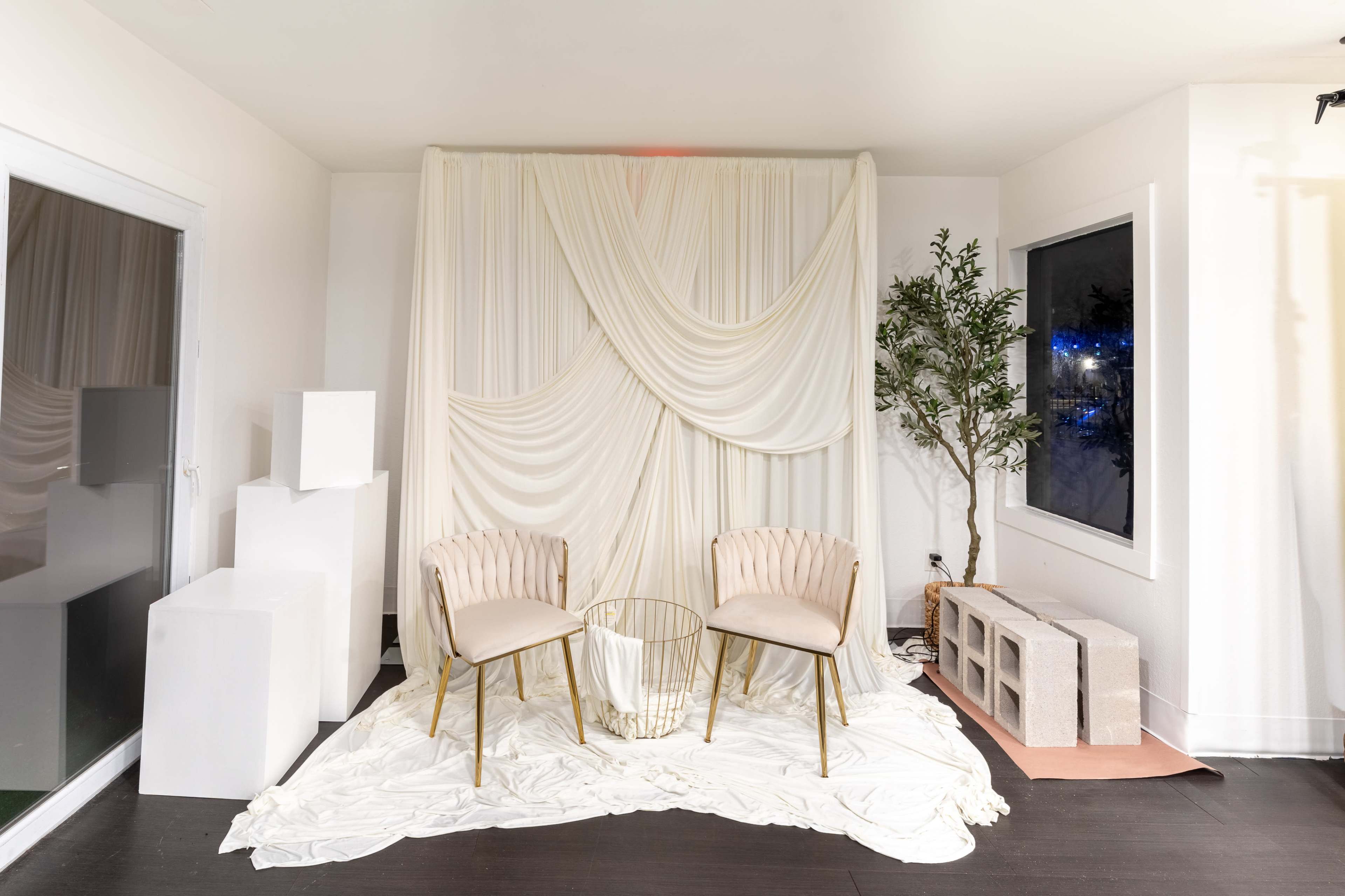 The image shows a minimalist seating area featuring two cream-colored chairs, a circular glass table, and a draped white backdrop with greenery beside a window.