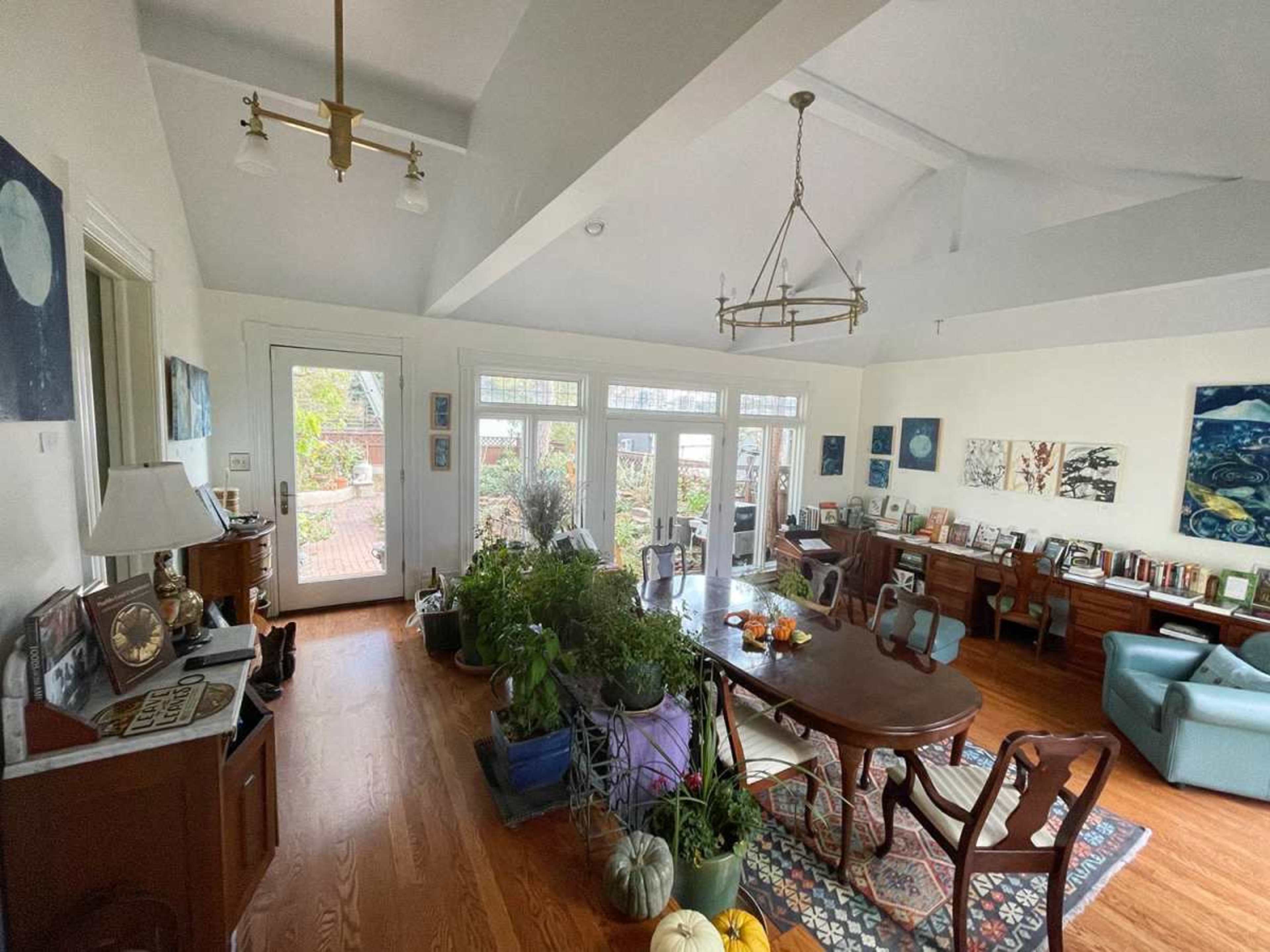 The image shows a bright indoor space with a dining table surrounded by chairs, bookshelves, and potted plants, leading to a patio with large windows.