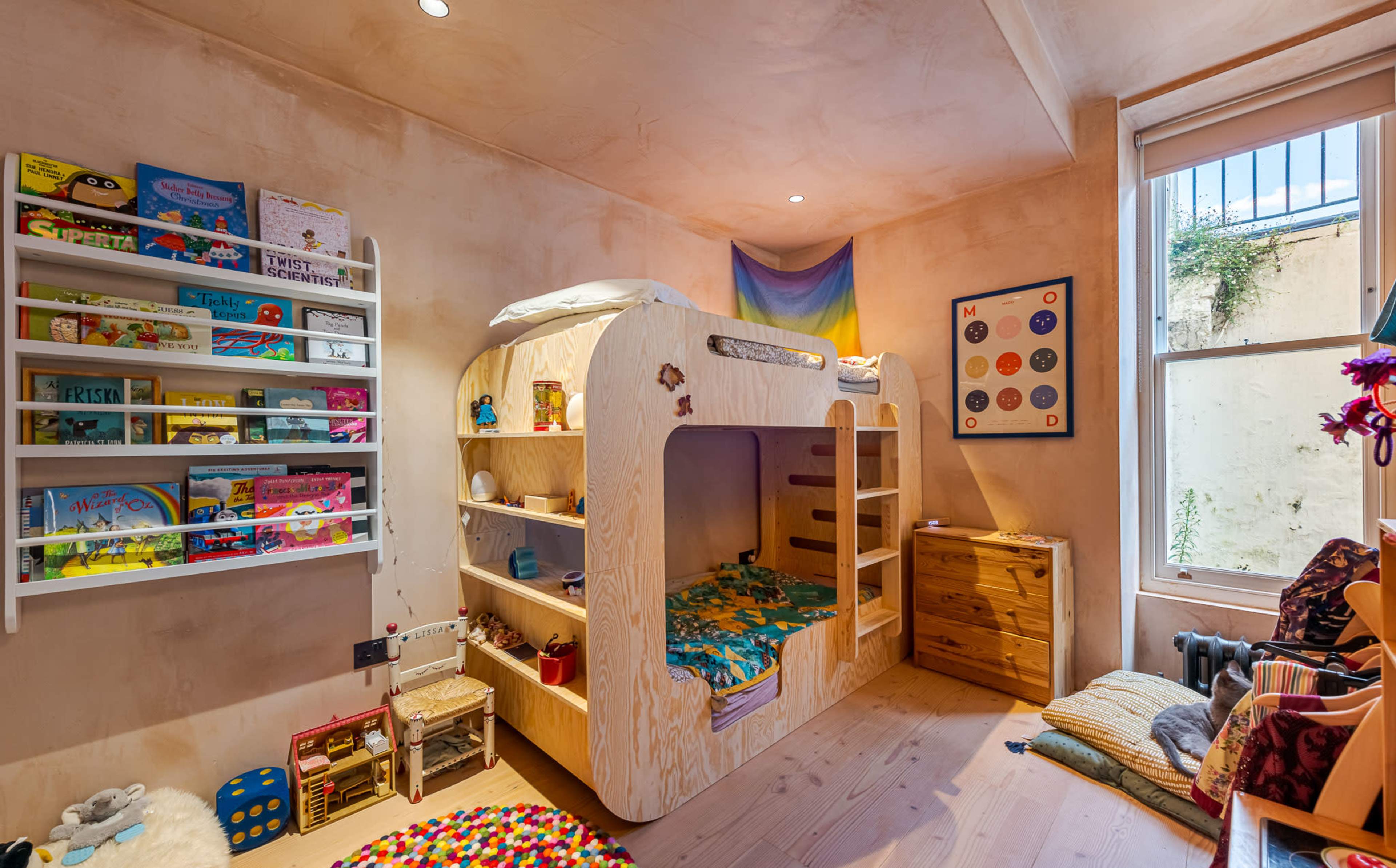 A children's bedroom featuring a wooden bunk bed structure with a colorful bedding set, shelves filled with books and toys, and a large window.