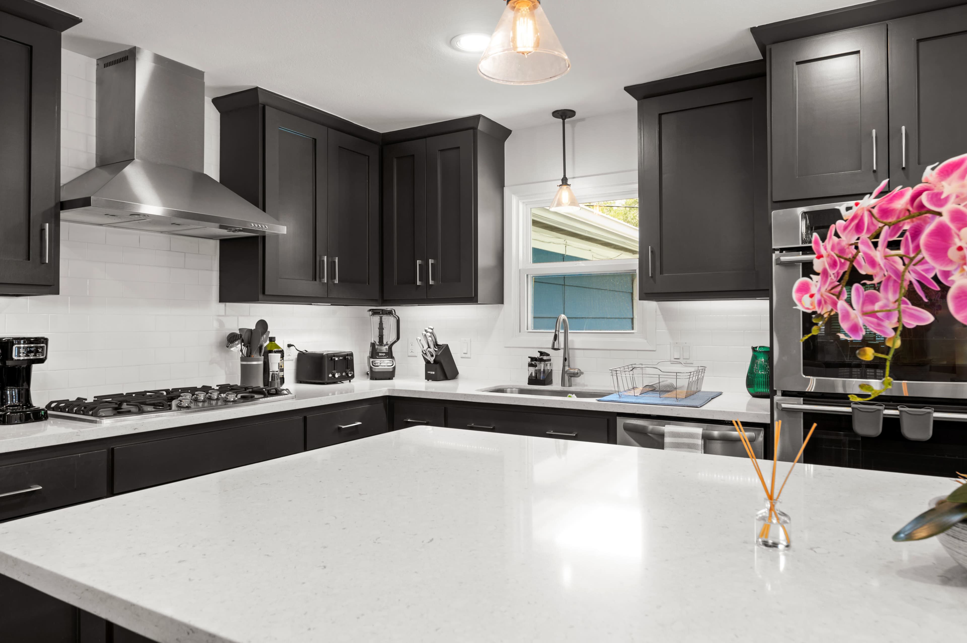 A modern kitchen features dark cabinetry, a large white countertop, and stainless steel appliances, with a decorative orchid on the counter.