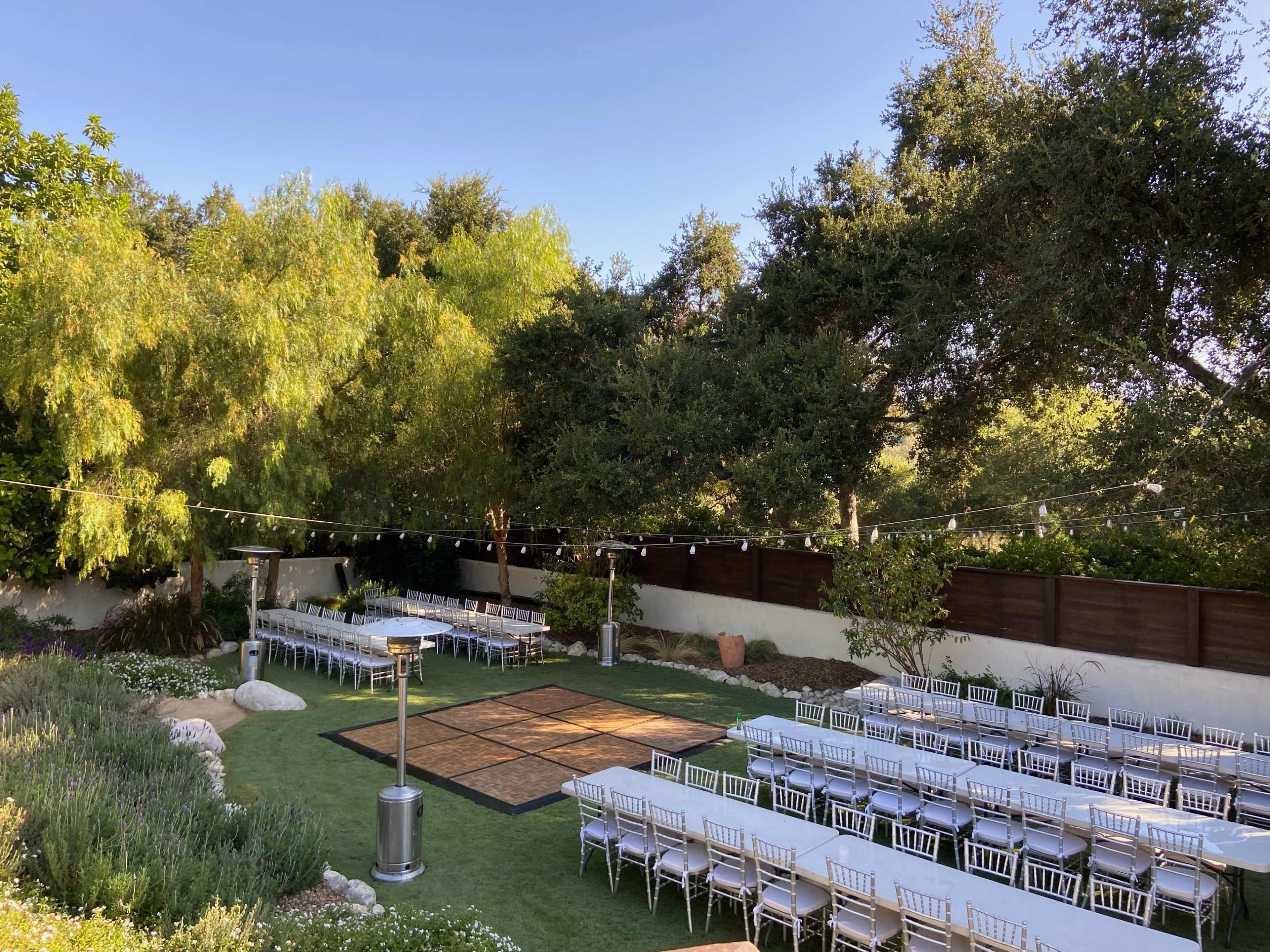 An outdoor event space with neatly arranged rows of tables and chairs set around a dance floor, surrounded by greenery and decorative lights.