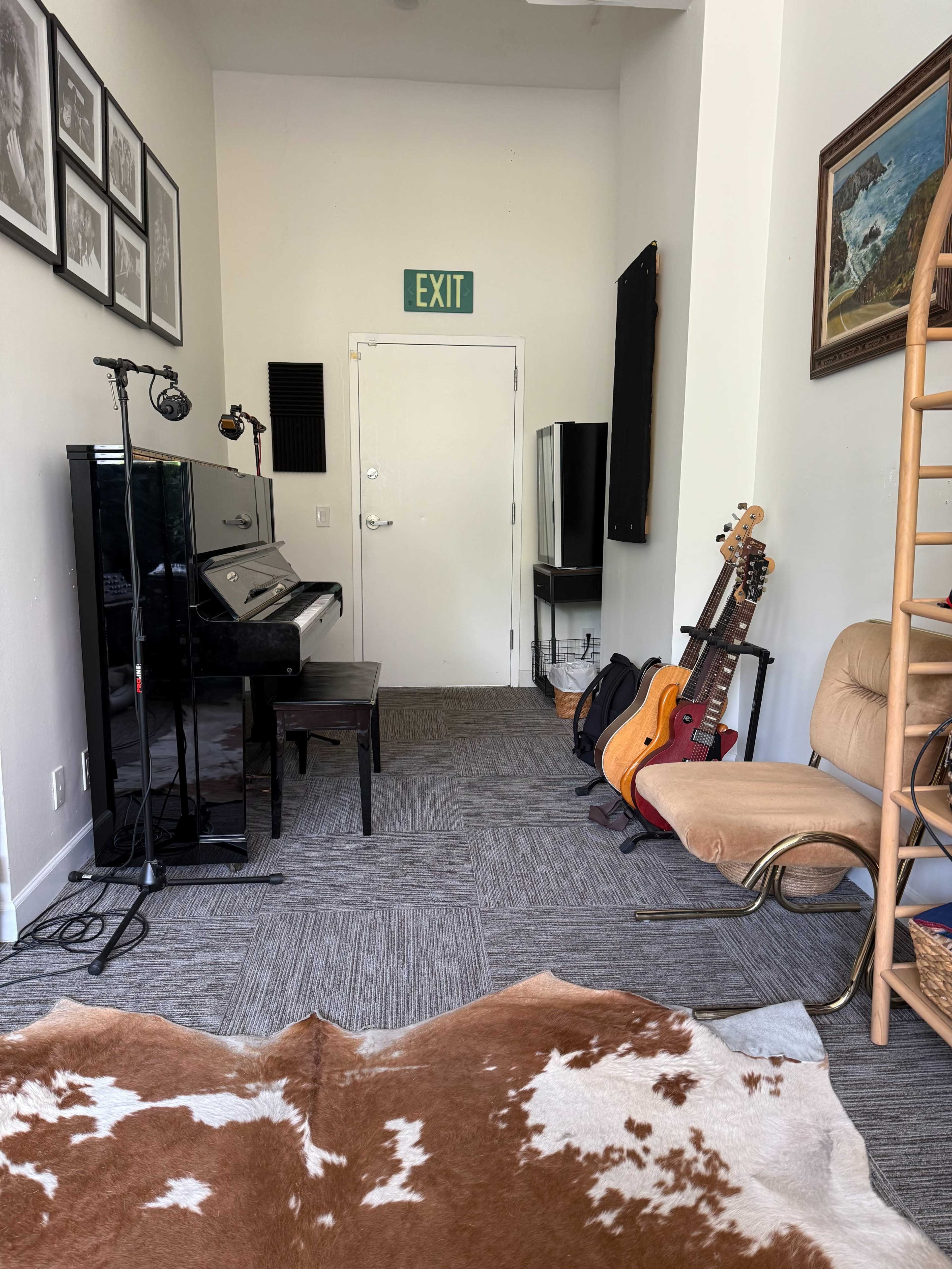 The image shows a narrow room with a black piano, guitars leaning against a chair, and a cowhide rug on the floor, leading to an exit door.