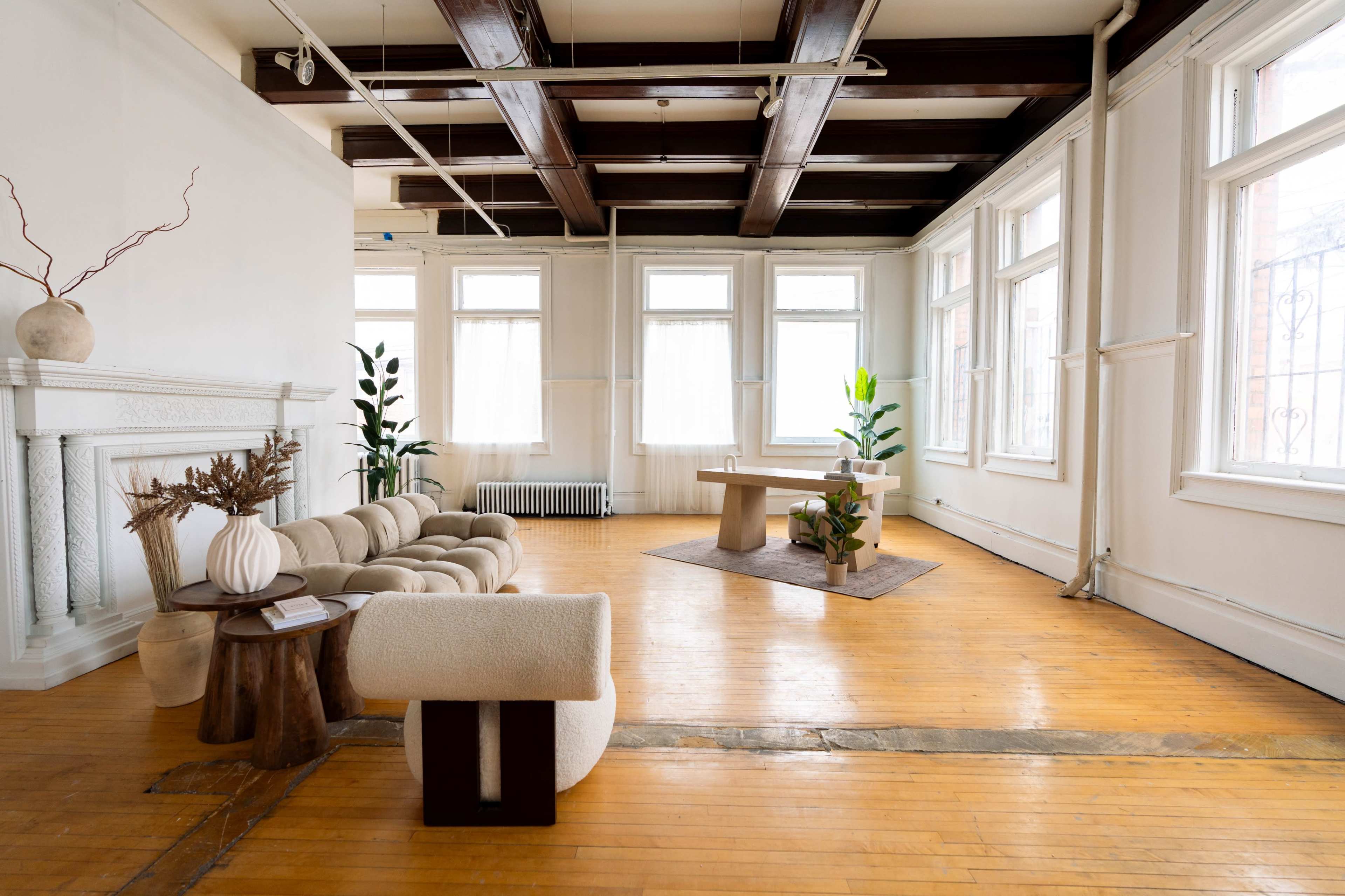 The image shows a spacious, minimalist room featuring a light wooden floor, large windows, and modern furniture including a curved sofa, a wooden table, and decorative plants.