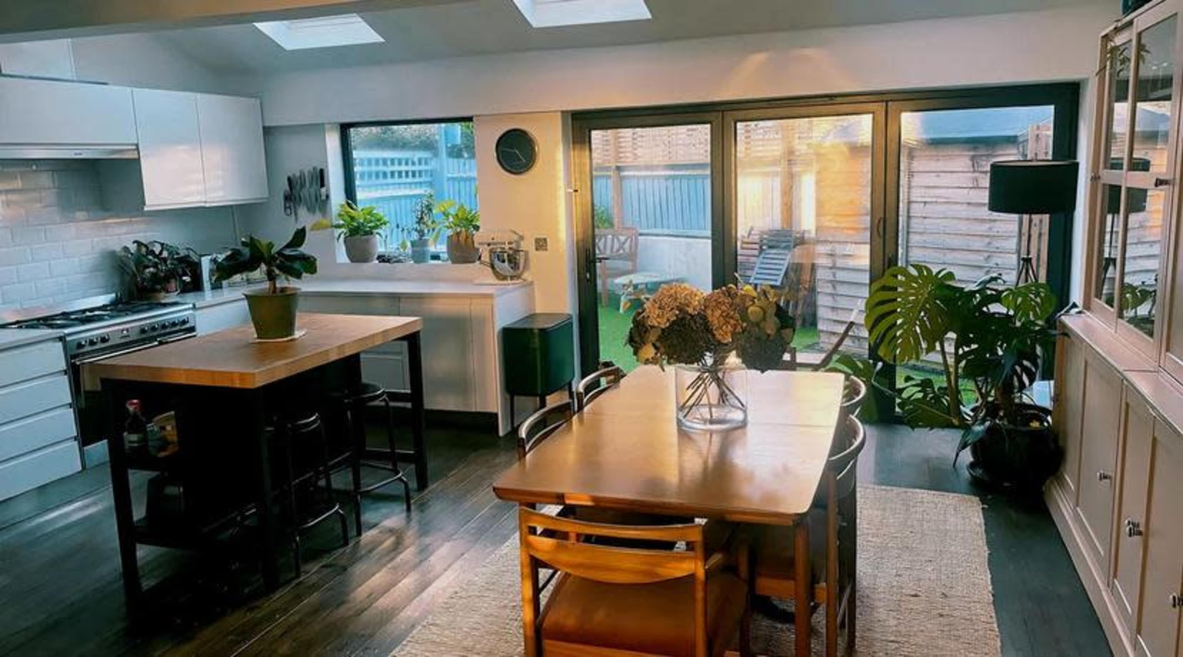 A modern kitchen and dining area features a wooden table with chairs, plants on the counters, and large windows that provide natural light.