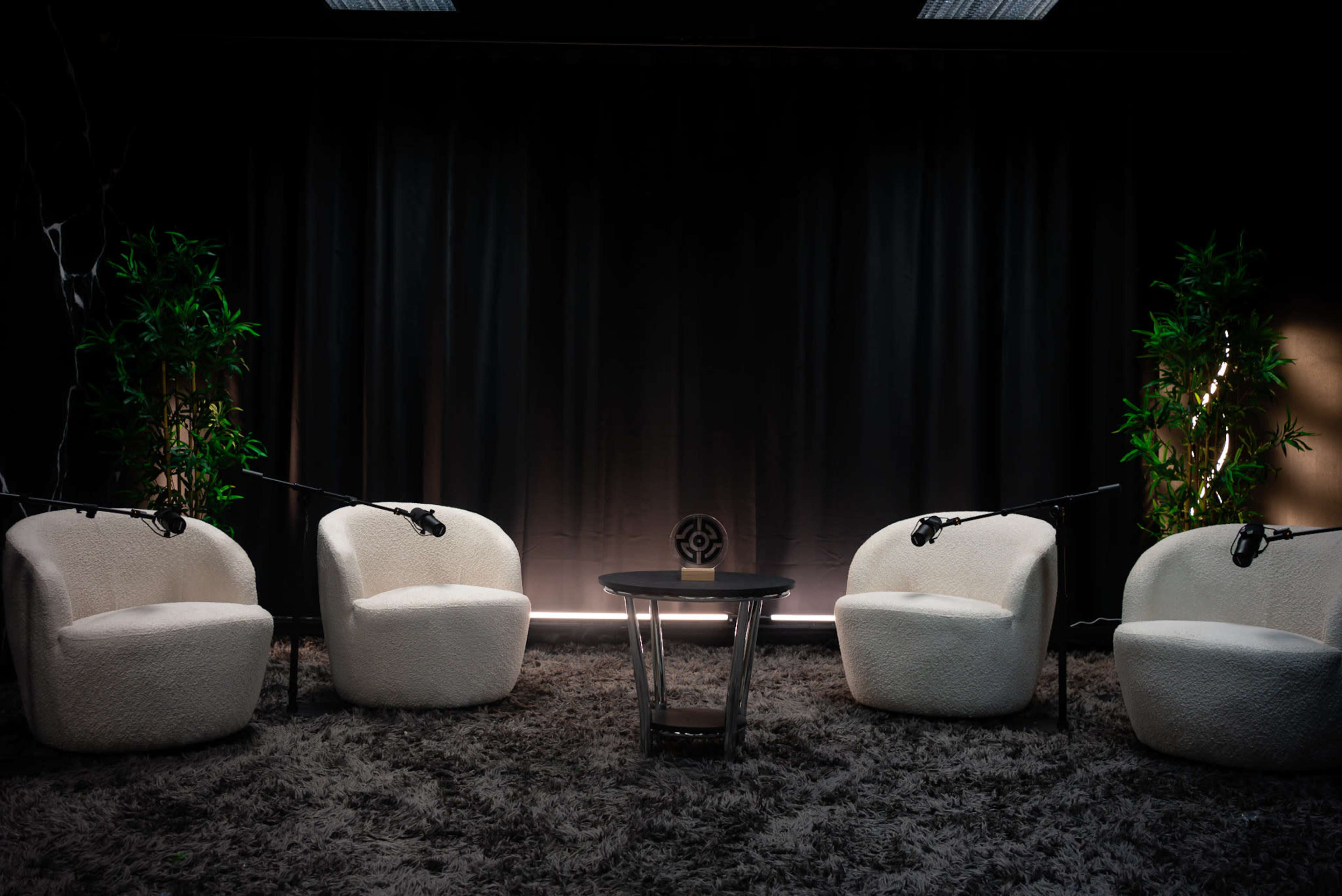 The image shows a small, cozy studio setup with four white, plush chairs arranged around a central table on a textured gray rug, with two green plants flanking the backdrop and dark curtains.