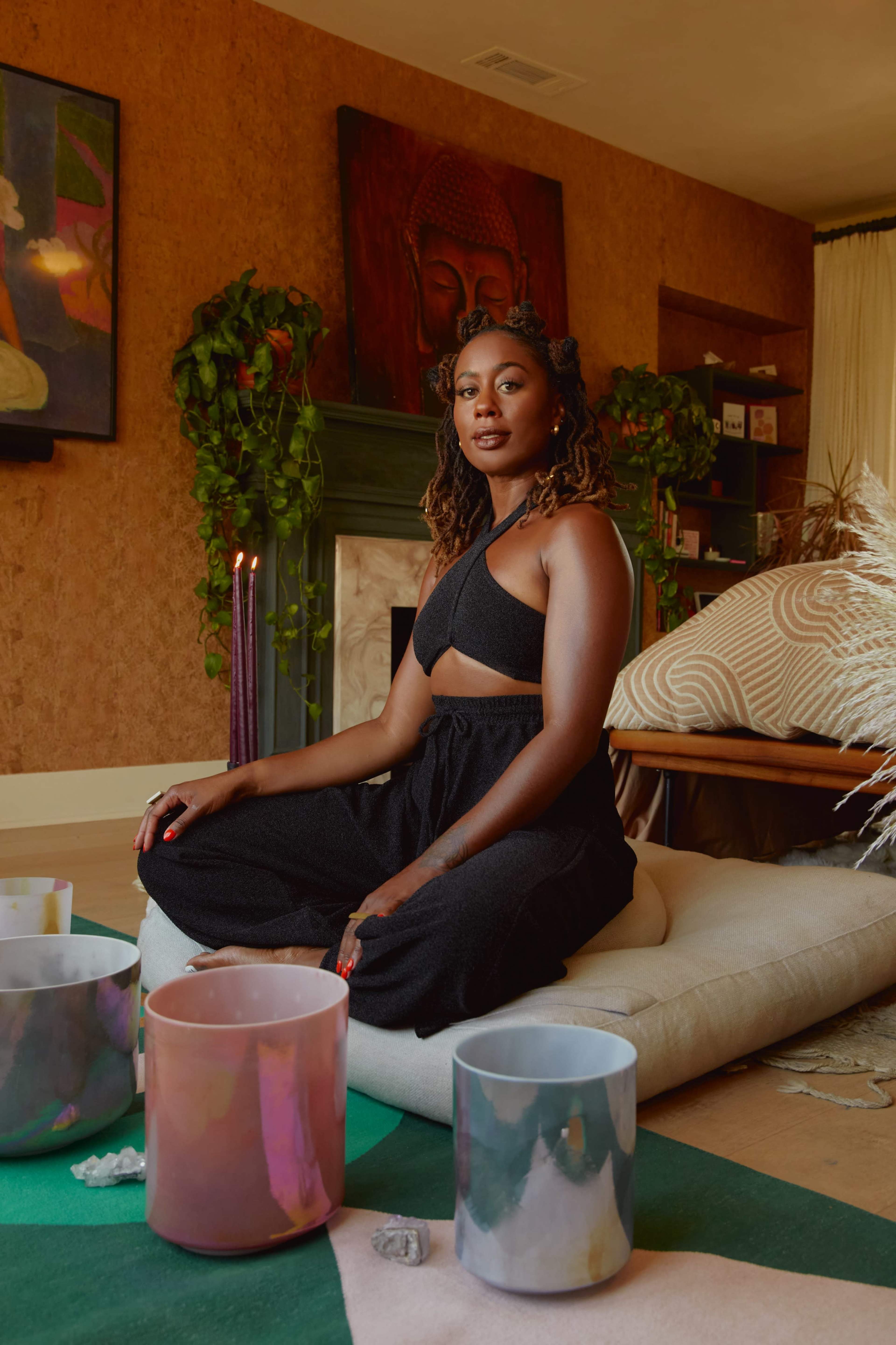 A woman sits cross-legged on a cushion in a warmly decorated room with plants, candles, and crystal singing bowls arranged in front of her.