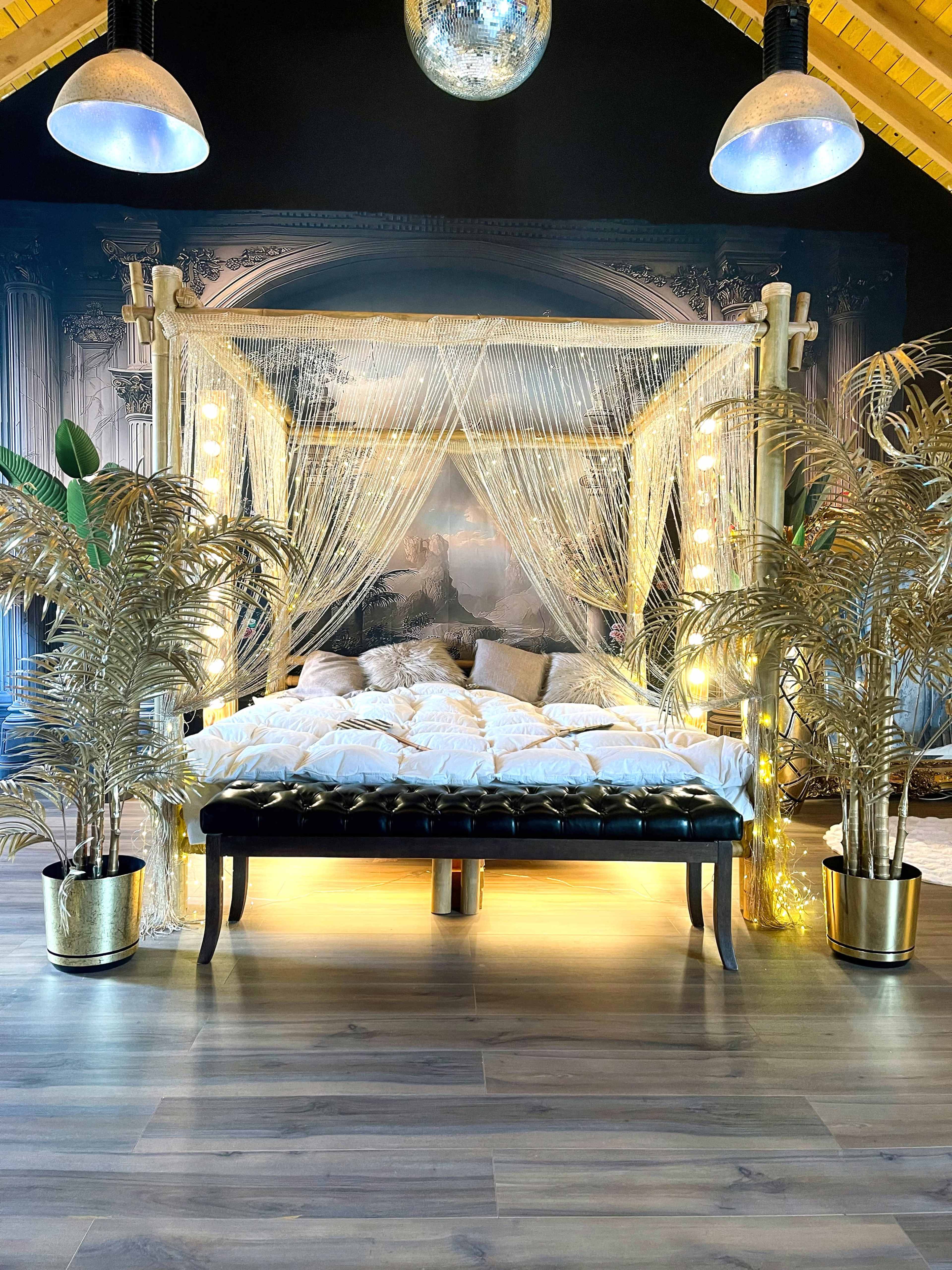 A rustic wooden bed with draped sheer curtains is surrounded by decorative plants and dim lighting in a cozy, well-decorated room.