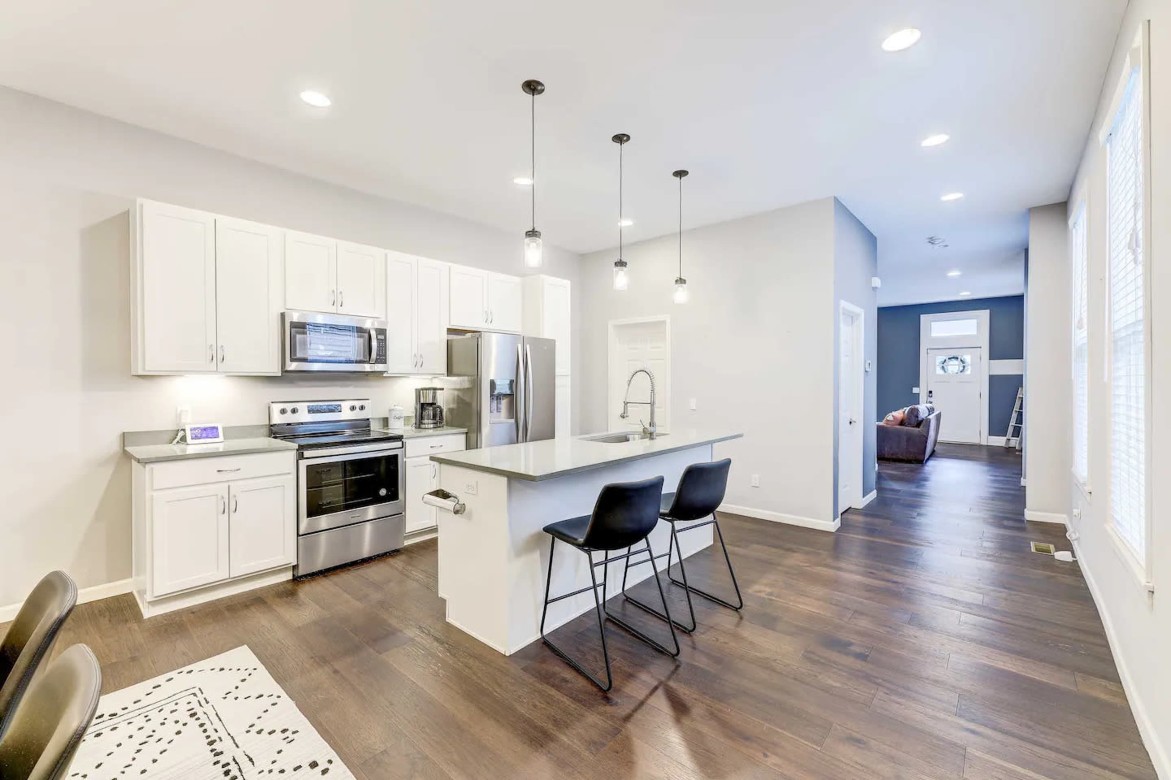 A modern kitchen with white cabinets, stainless steel appliances, and a central island with three black bar stools, leading into a well-lit living area.
