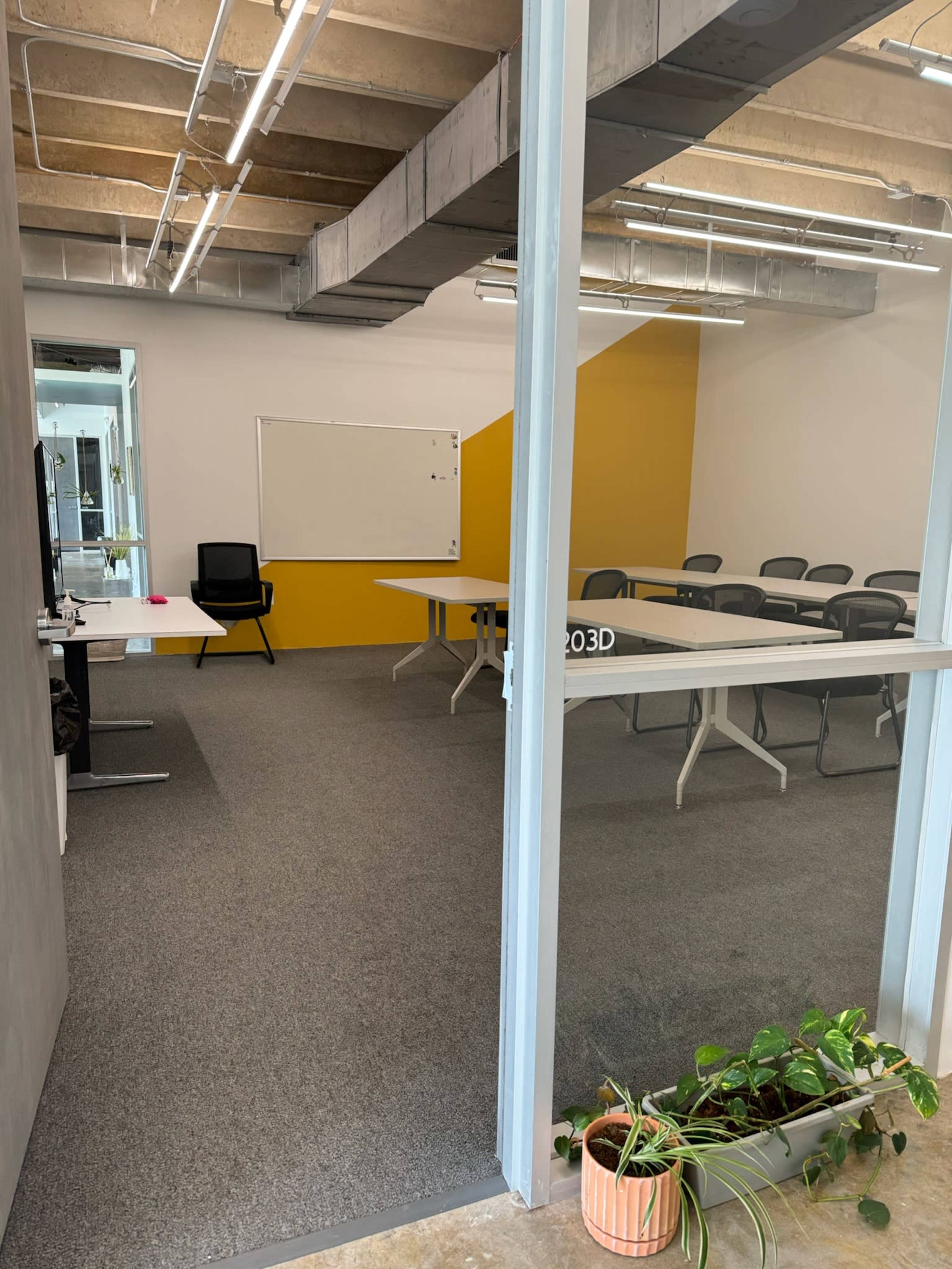 The image shows a modern conference room with a long table, several black chairs, a whiteboard, and a glass door opening to the space.