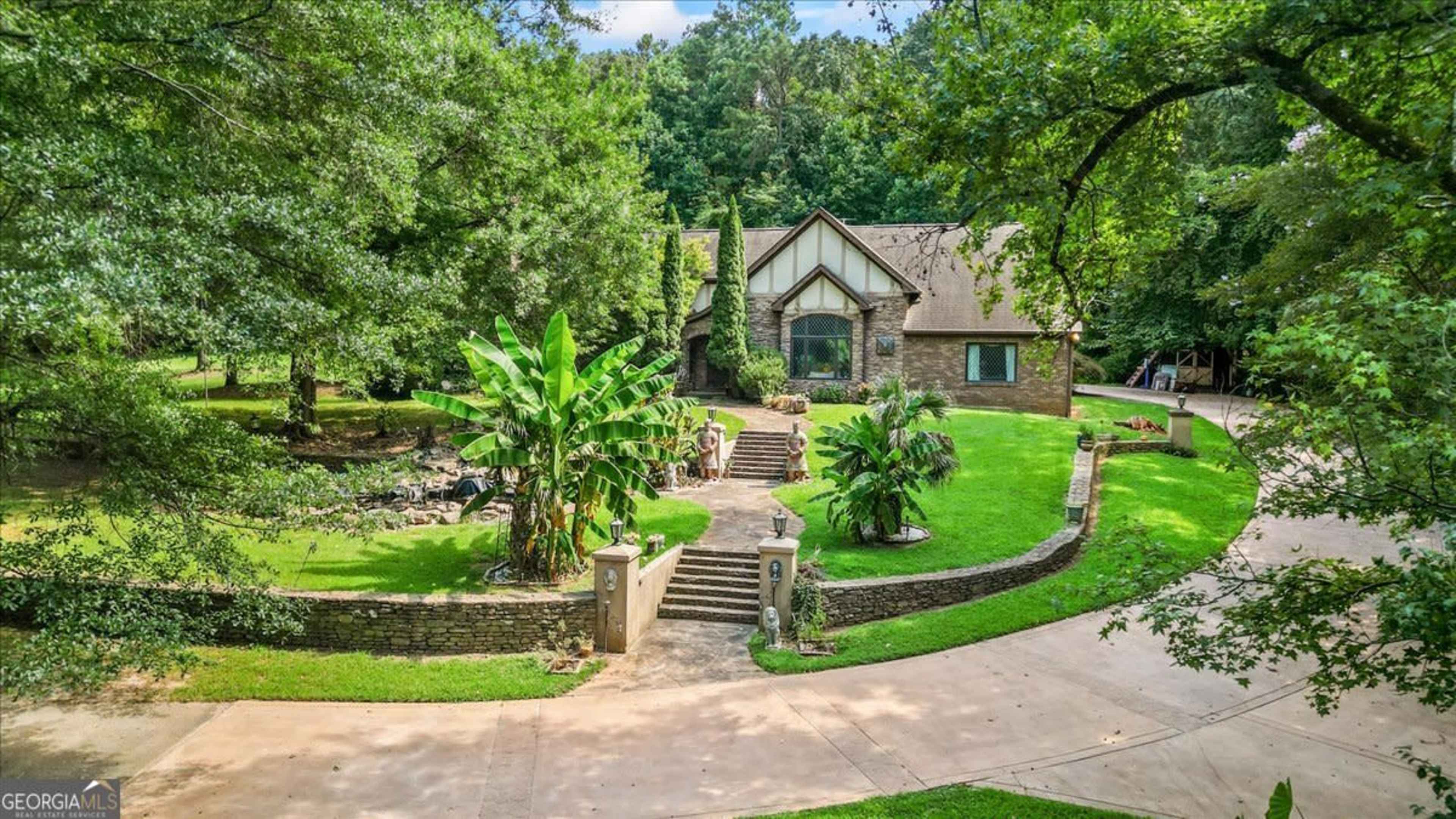 A large, two-story house with a stone facade and a landscaped yard featuring palm trees and a circular driveway is surrounded by dense greenery.
