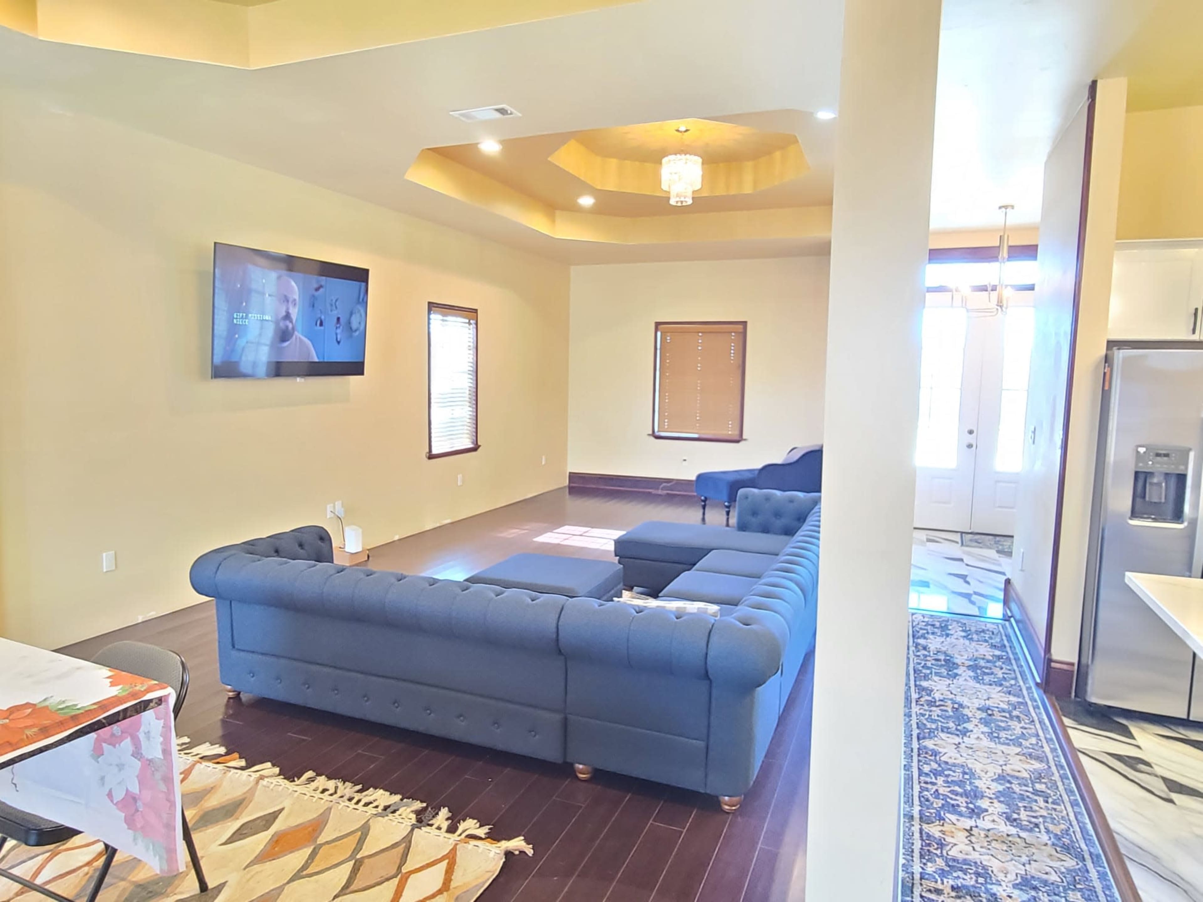 A spacious living room featuring a large blue sectional couch, a television mounted on the wall, and natural light coming through several windows.