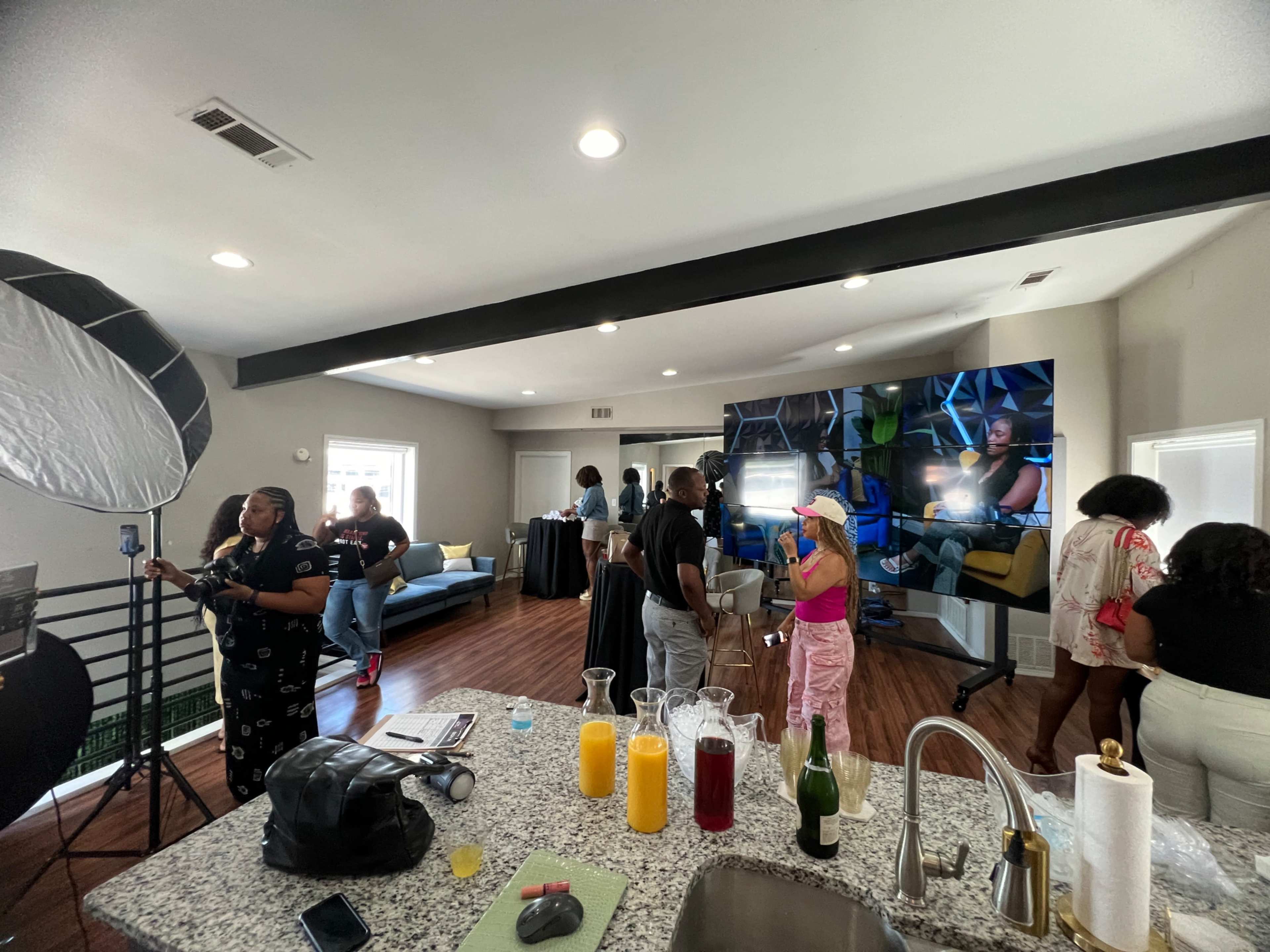 A group of people is gathered in a modern living space with a large screen displaying images, while others prepare drinks and snacks on a kitchen countertop.