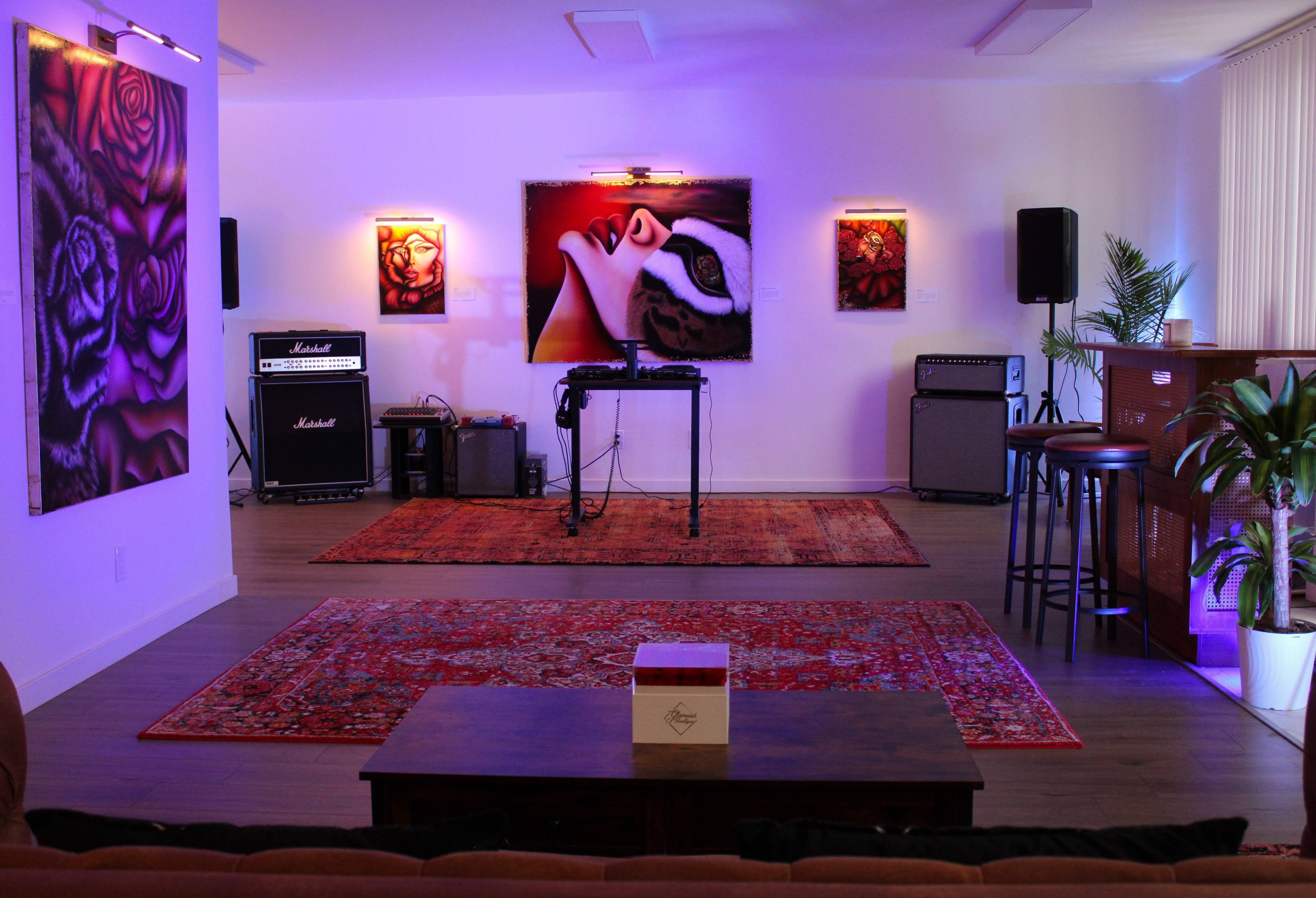 The room features a cozy lounge area with a wooden coffee table, surrounded by vibrant artwork, a sound system, and a bar counter, all illuminated by soft purple lighting.