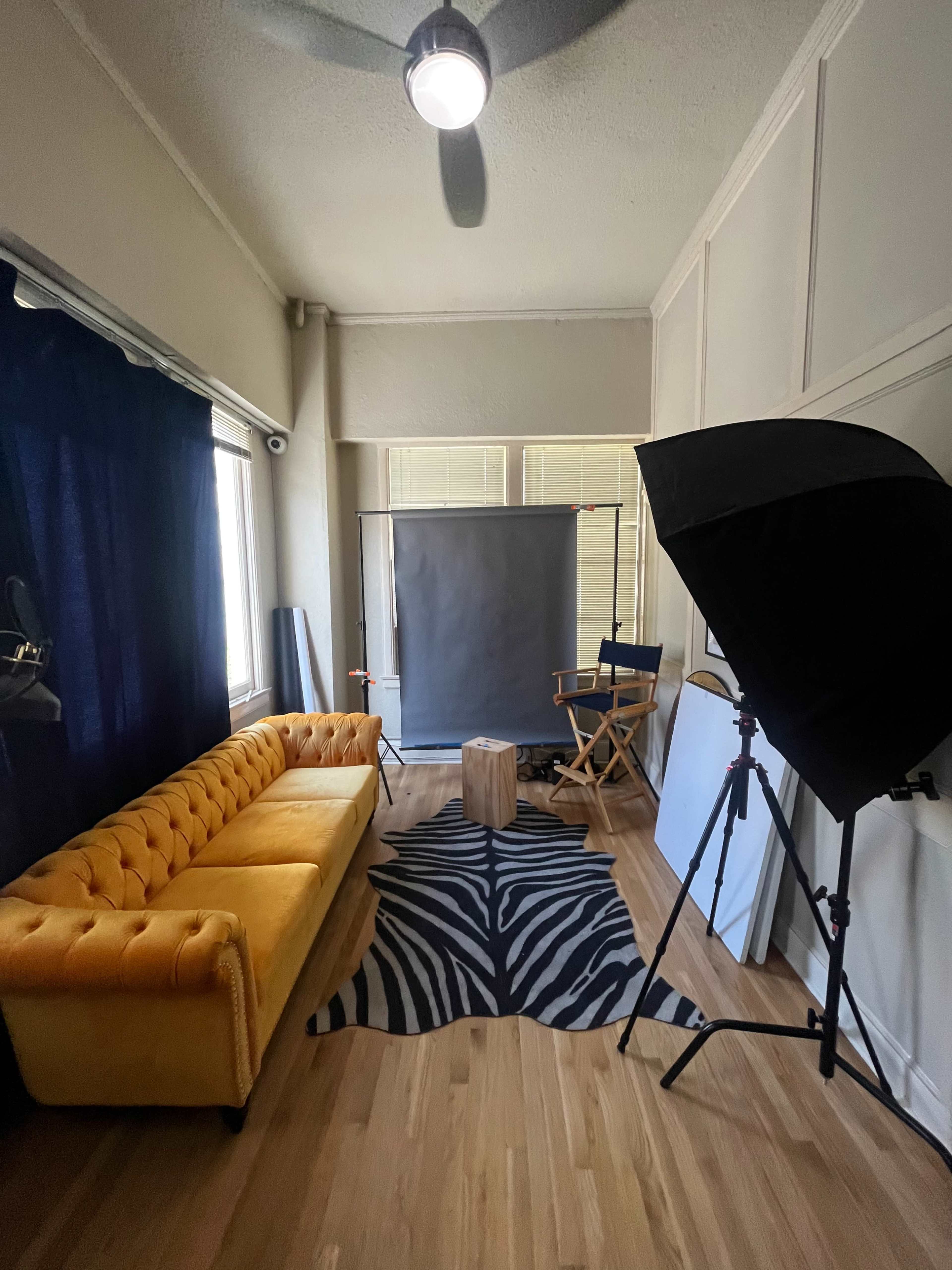 The image shows a photography studio with a yellow couch, a zebra-print rug, a wooden stool, a backdrop setup, and a studio light on a stand.