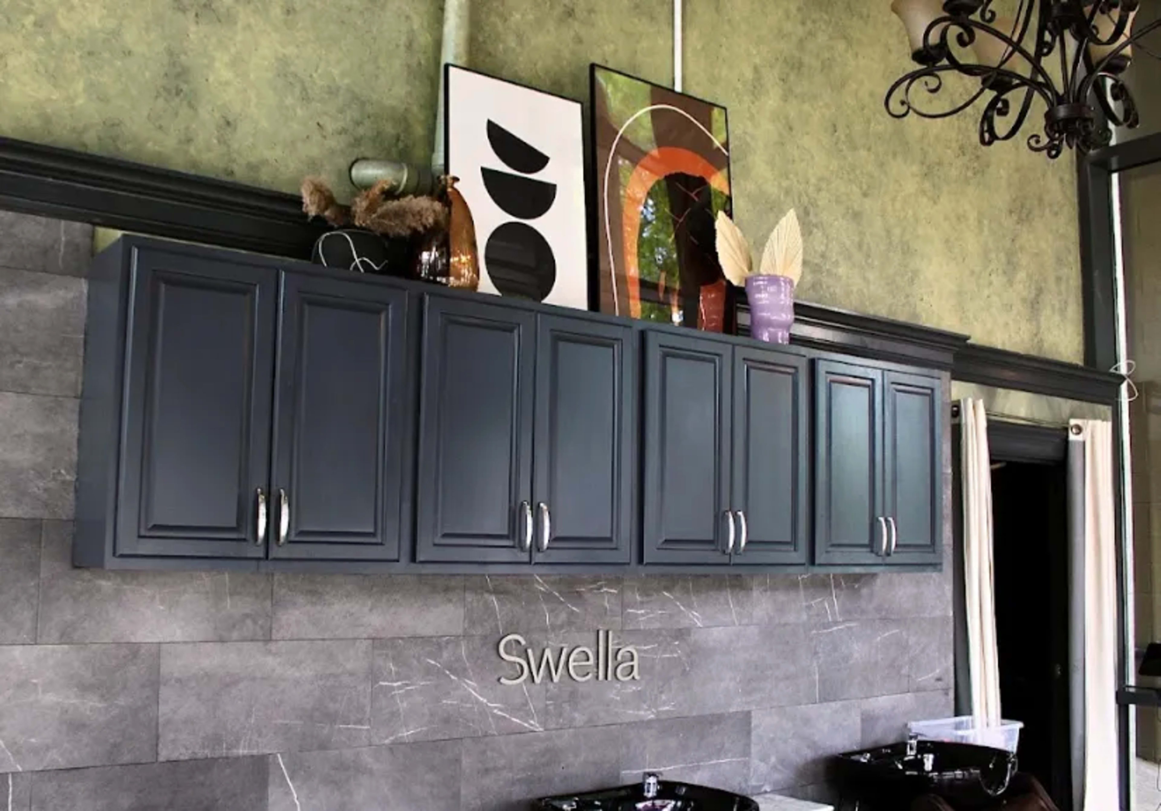 The image shows a modern kitchen with dark cabinetry above a countertop, featuring decorative artwork and a plant arrangement on top.