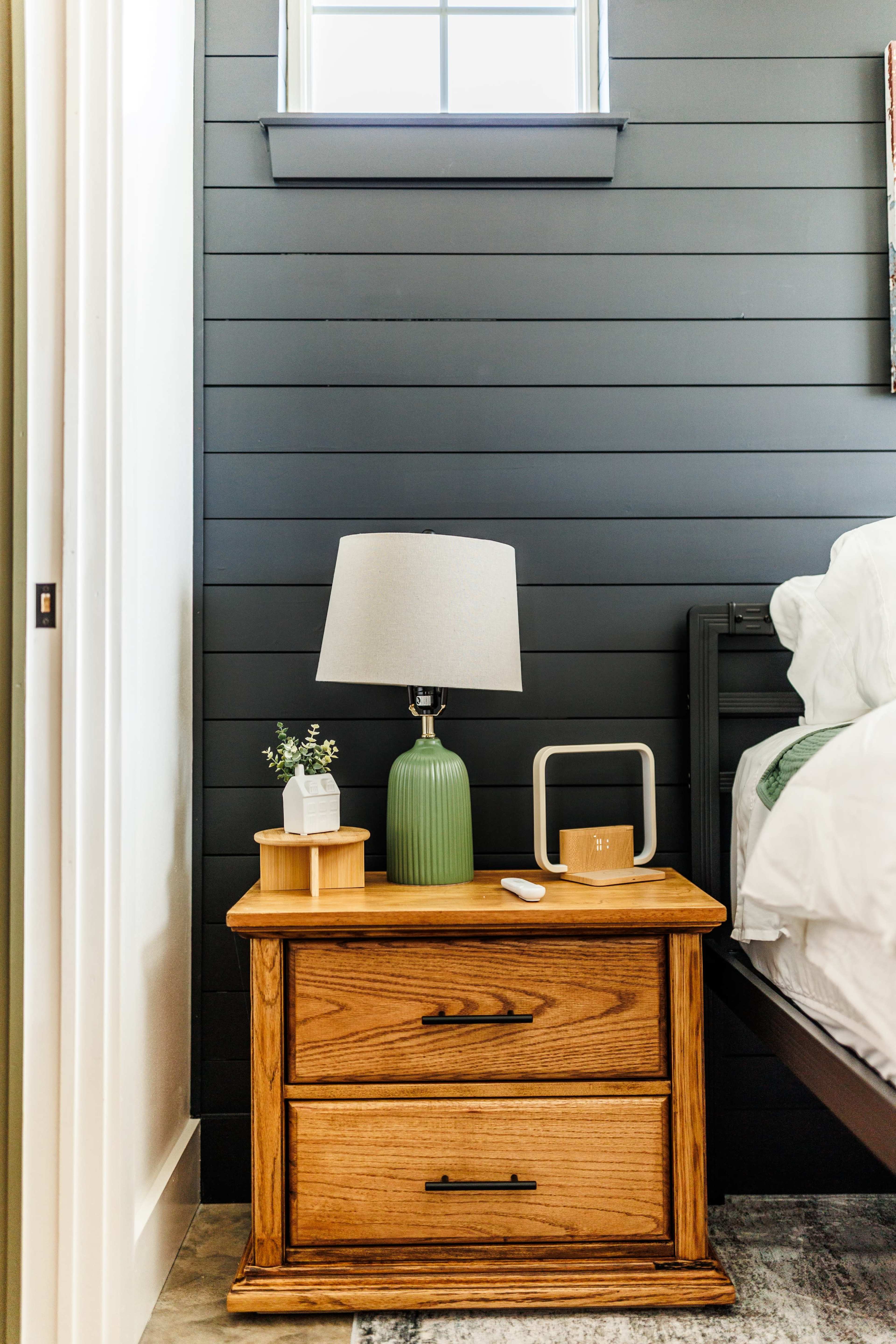 The image shows a wooden bedside table with two drawers, a green lamp, a small plant, and a white object, placed next to a bed against a dark paneled wall.