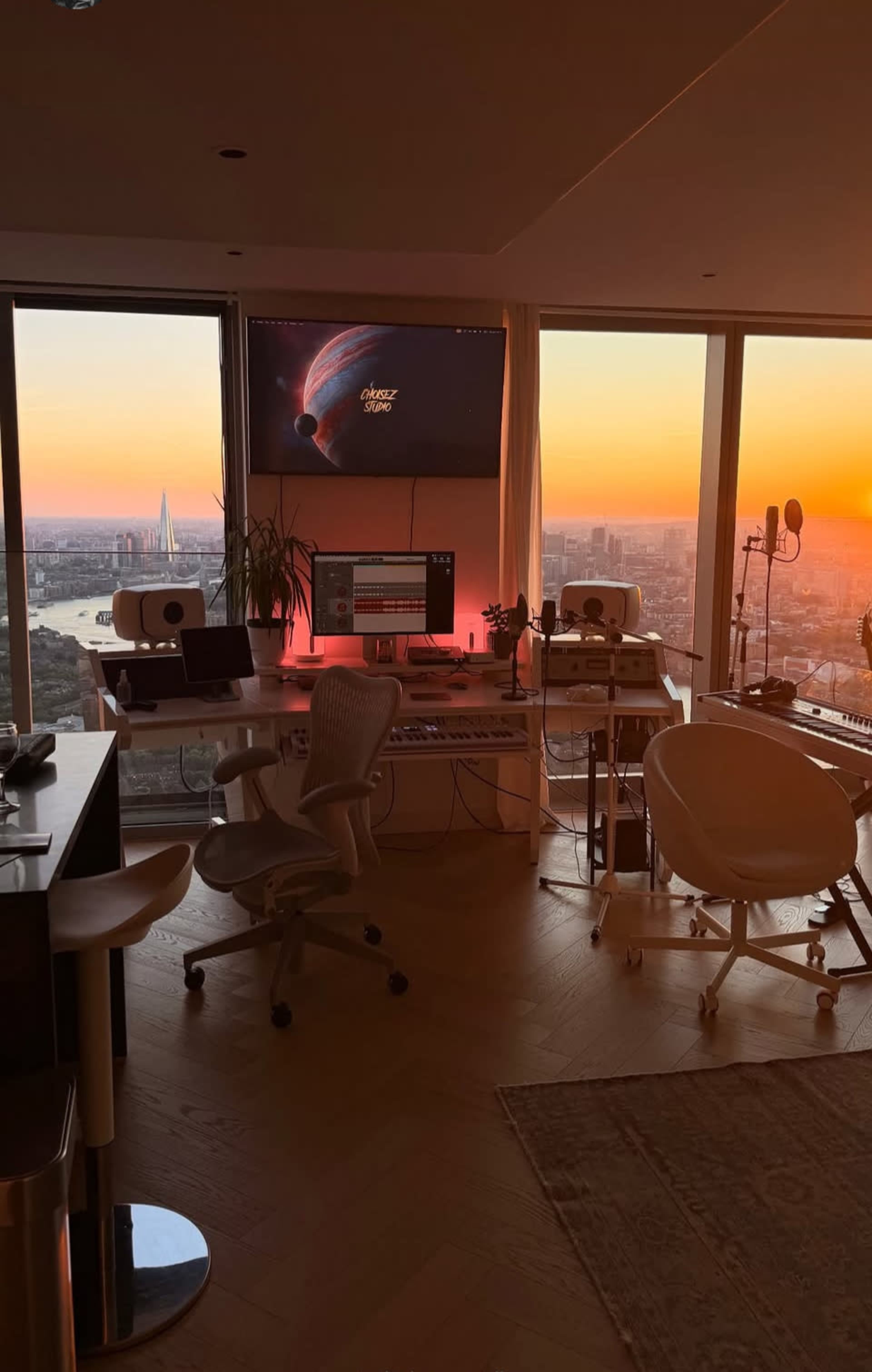 A modern workspace is illuminated by sunset, featuring a large monitor, audio equipment, and stylish furniture near a window overlooking a city skyline.