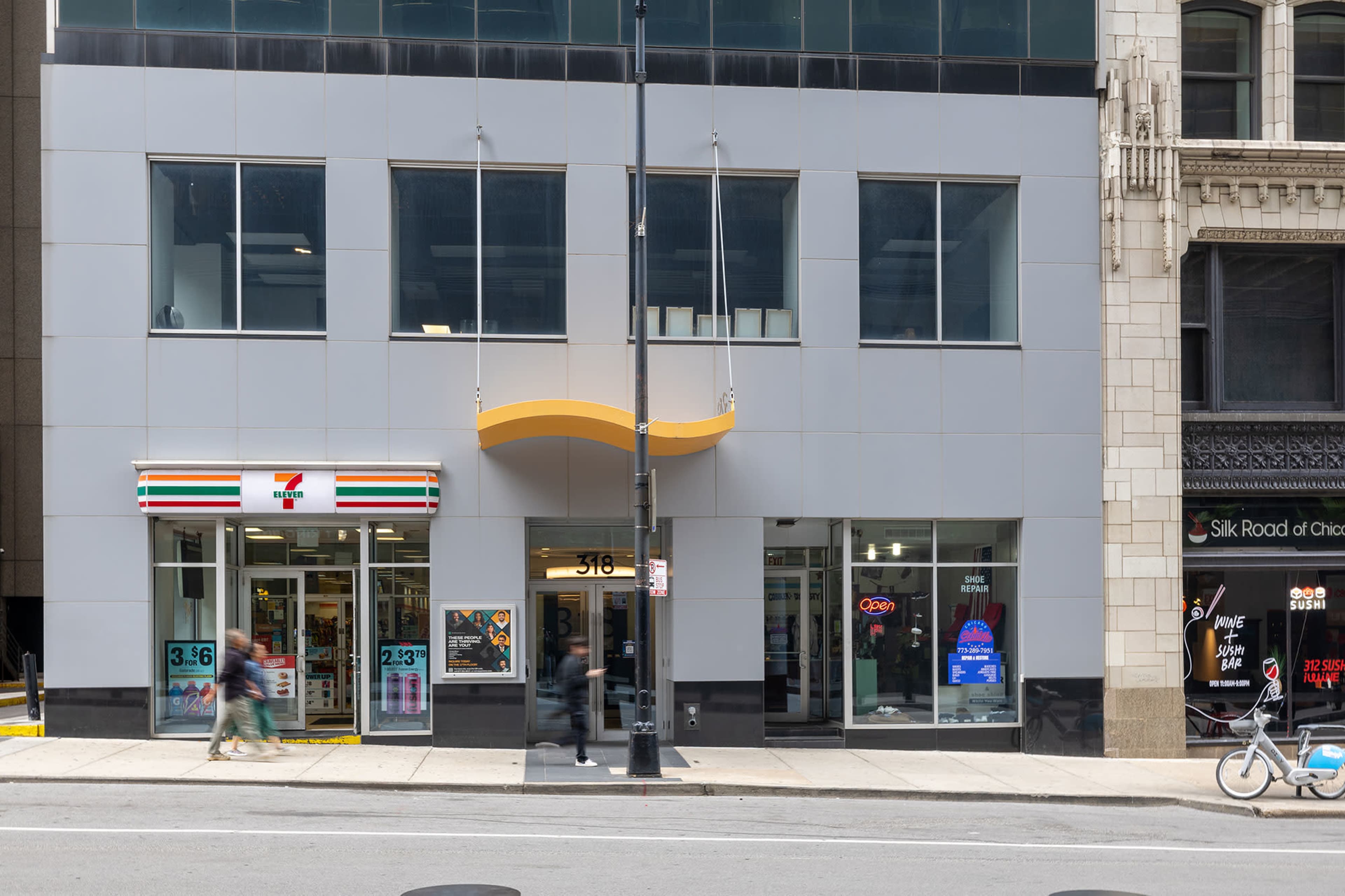 The image shows the exterior of a modern building featuring a 7-Eleven storefront on the left and a business entrance in the center, with large glass windows.