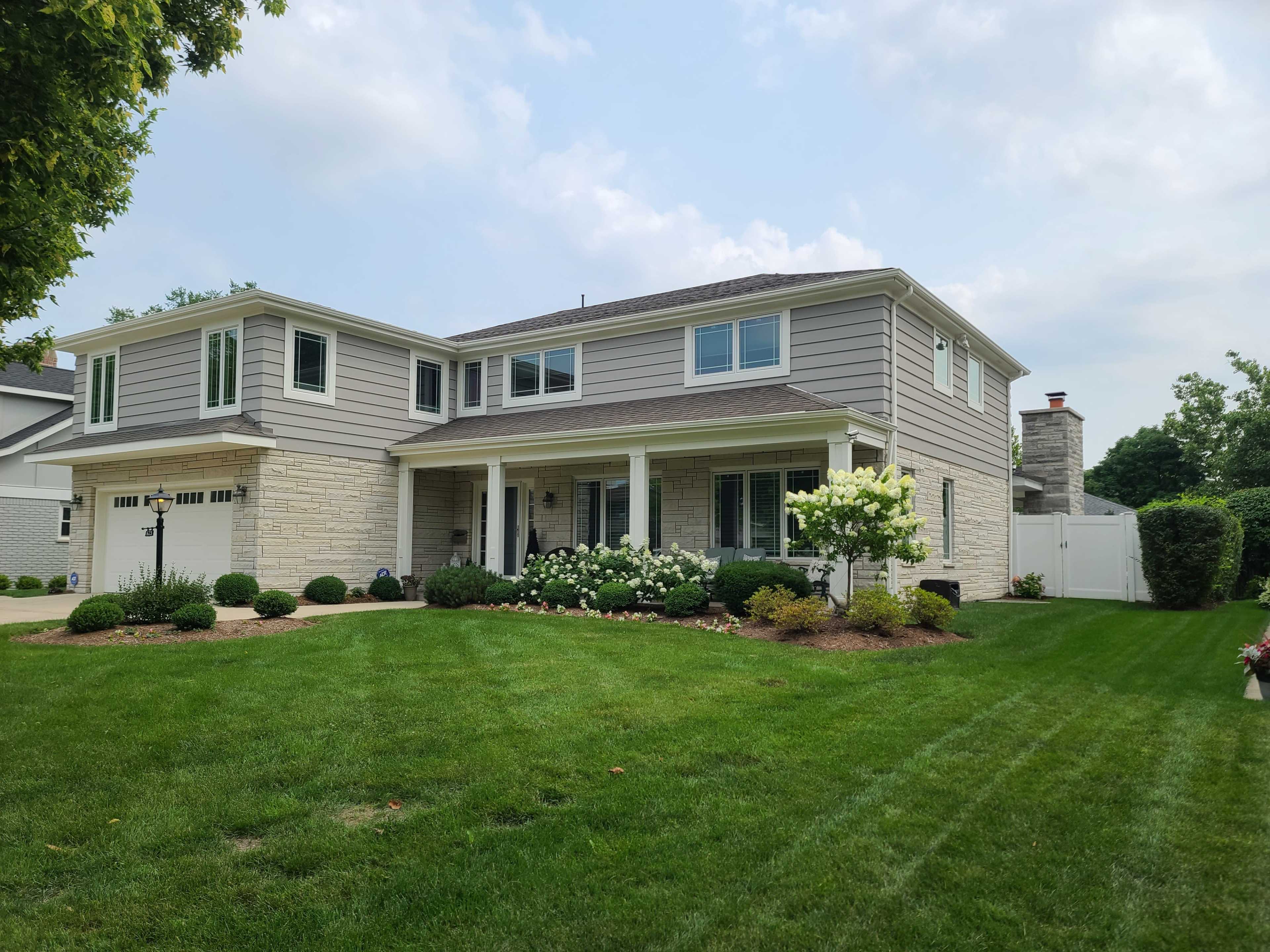 A two-story gray house with cream-colored stone accents and a well-maintained lawn featuring shrubs and flowers.