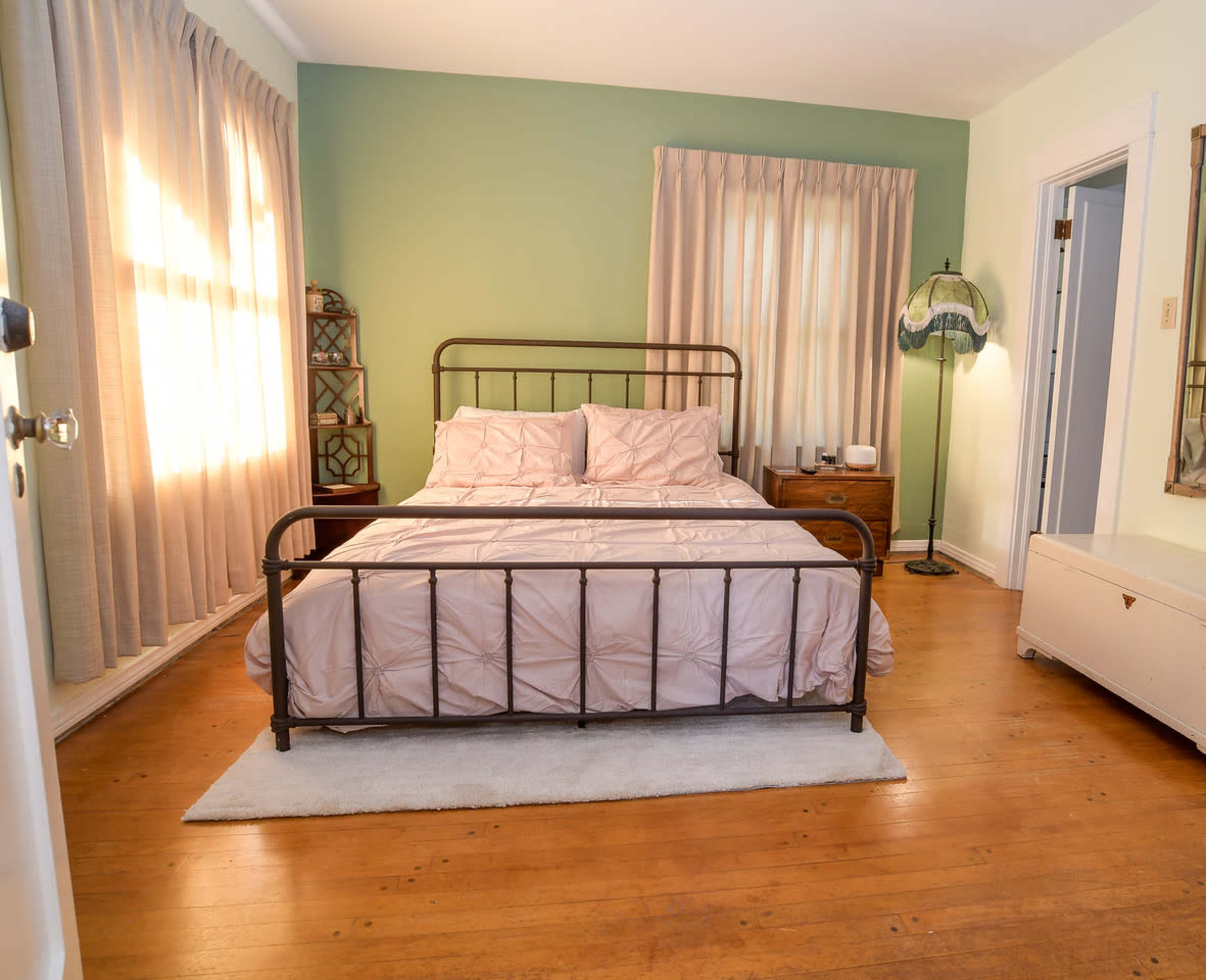 A bedroom with a metal bed frame, a light-colored comforter, and large windows draped with curtains.