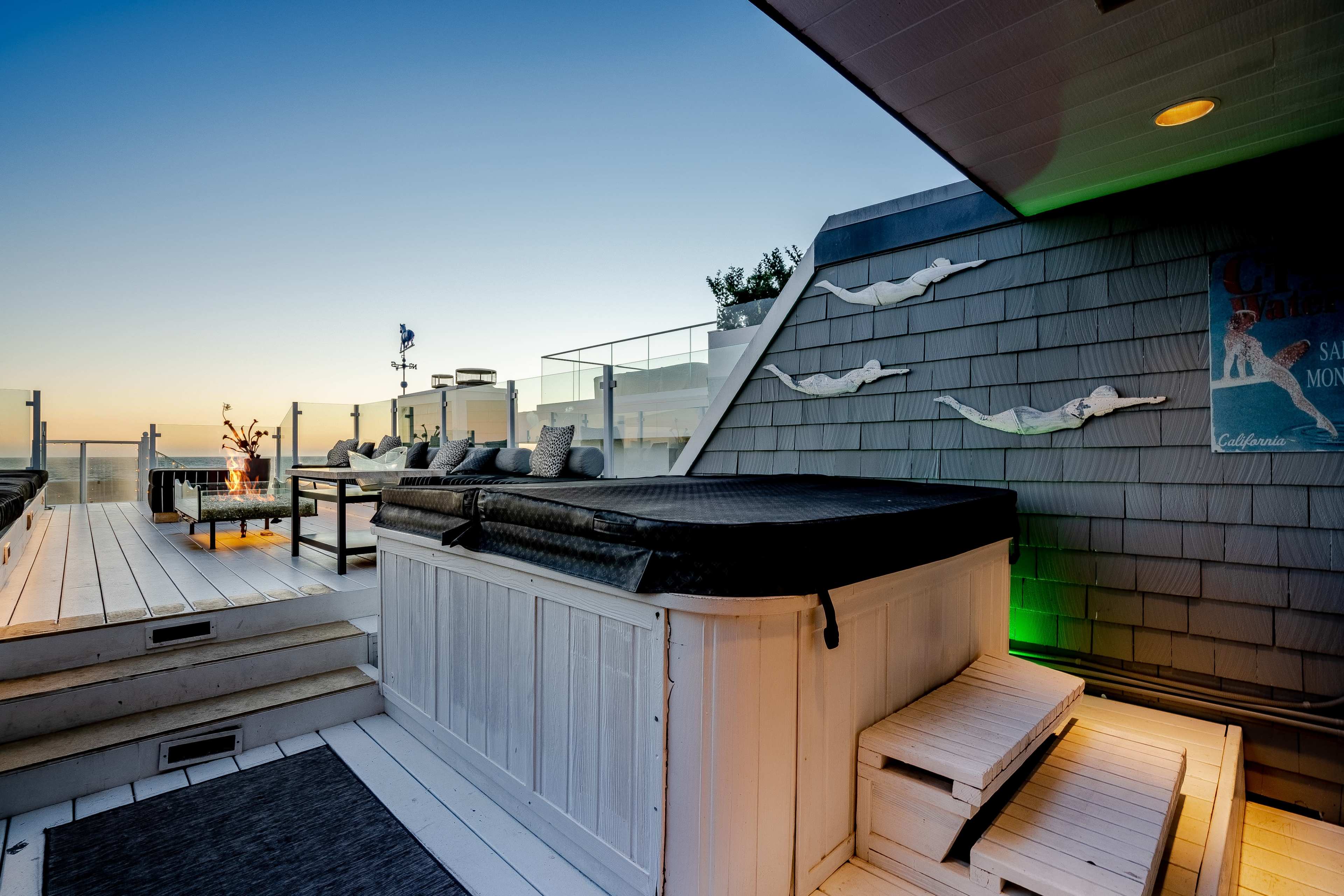 A rooftop deck with a hot tub, seating area, and an ocean view at sunset.