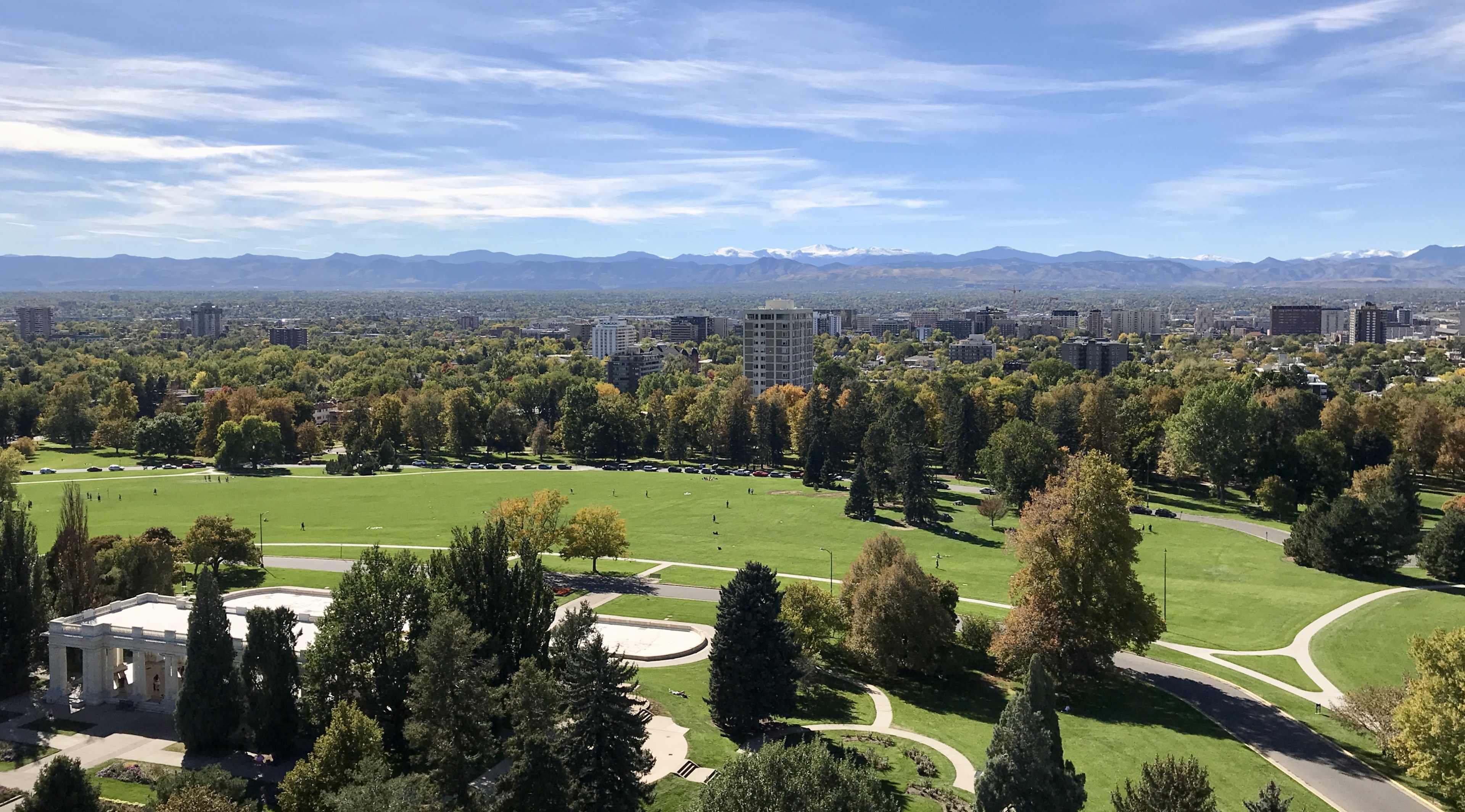 A sprawling city park with lush green lawns and trees, set against a backdrop of mountains and a clear blue sky.